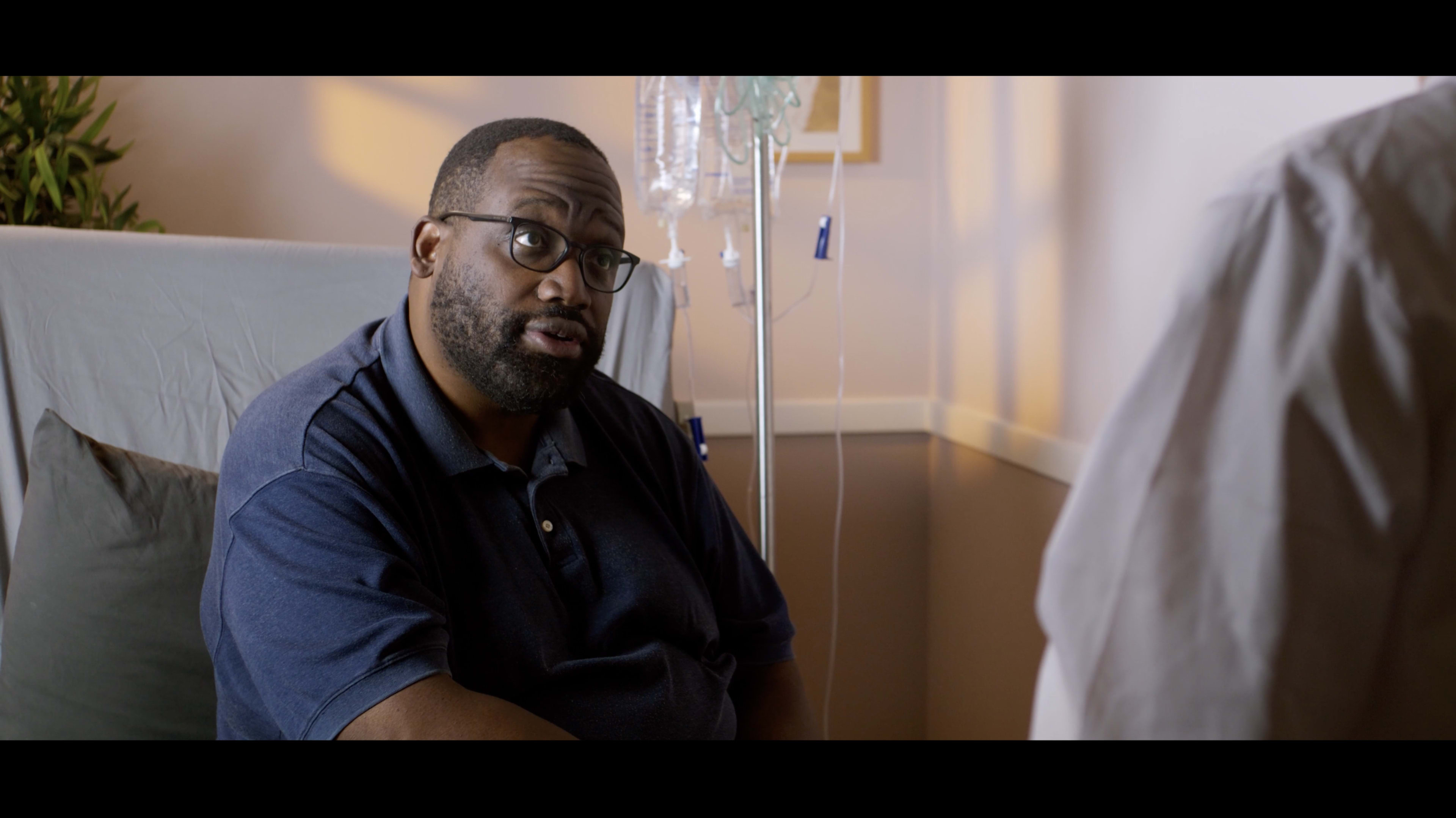 A man wearing glasses sits on a hospital bed while speaking with a medical professional.