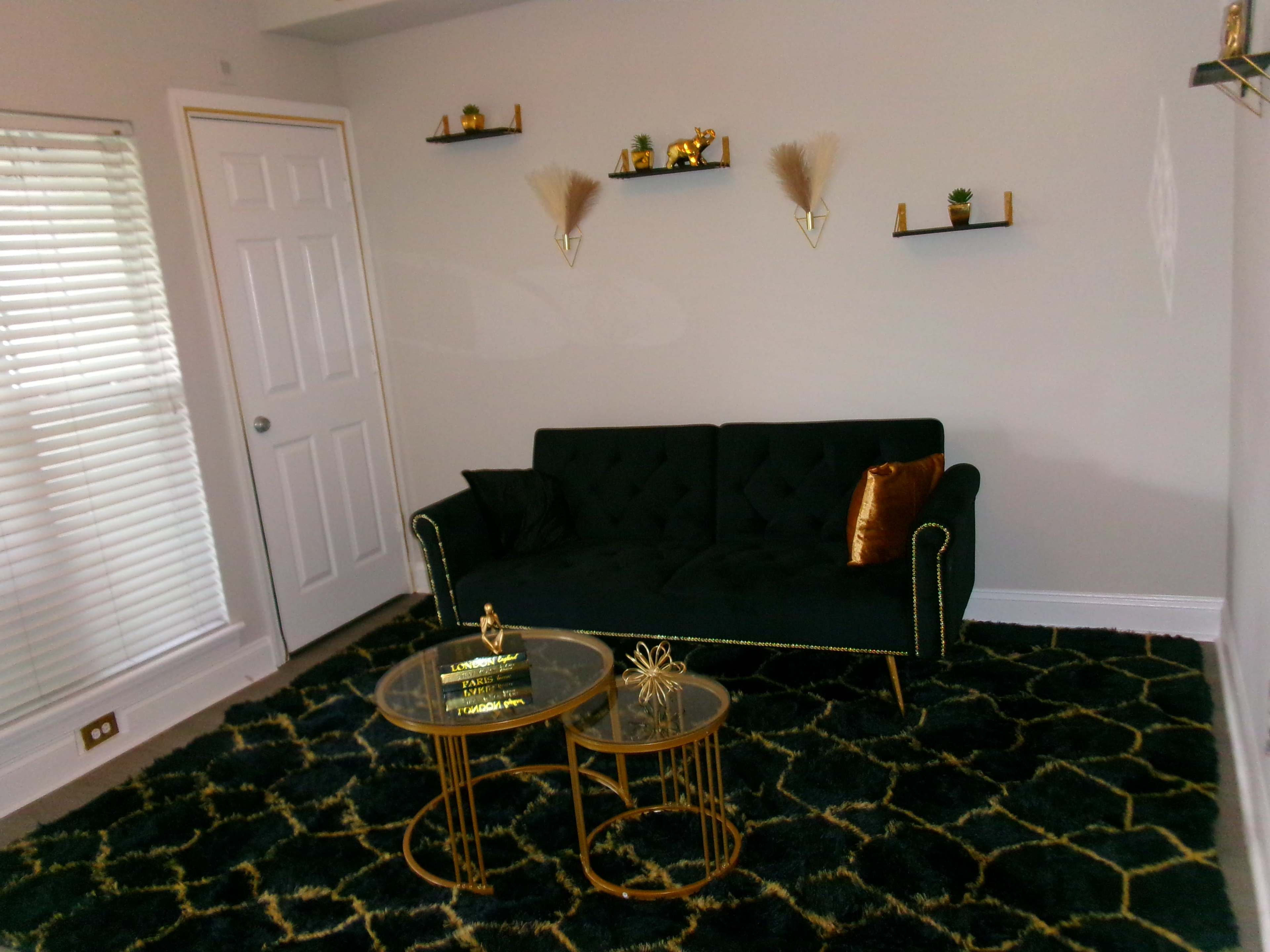 The image shows a living room with a dark green couch, two gold side tables, and decorative shelves on the wall.