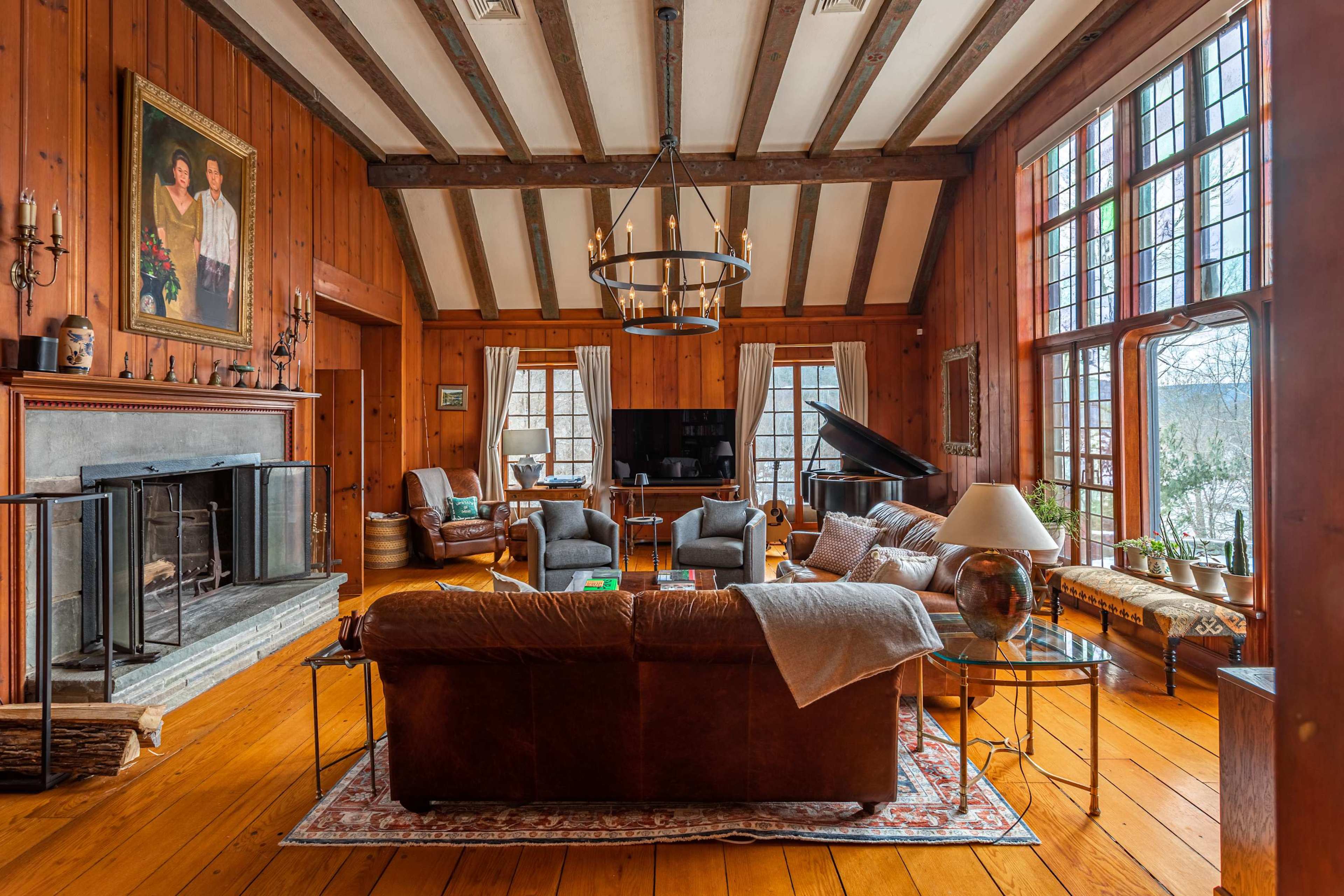 The image shows a cozy living room with wooden walls, a stone fireplace, large windows, and a mix of seating including a brown leather couch and armchairs facing a television and a grand piano.