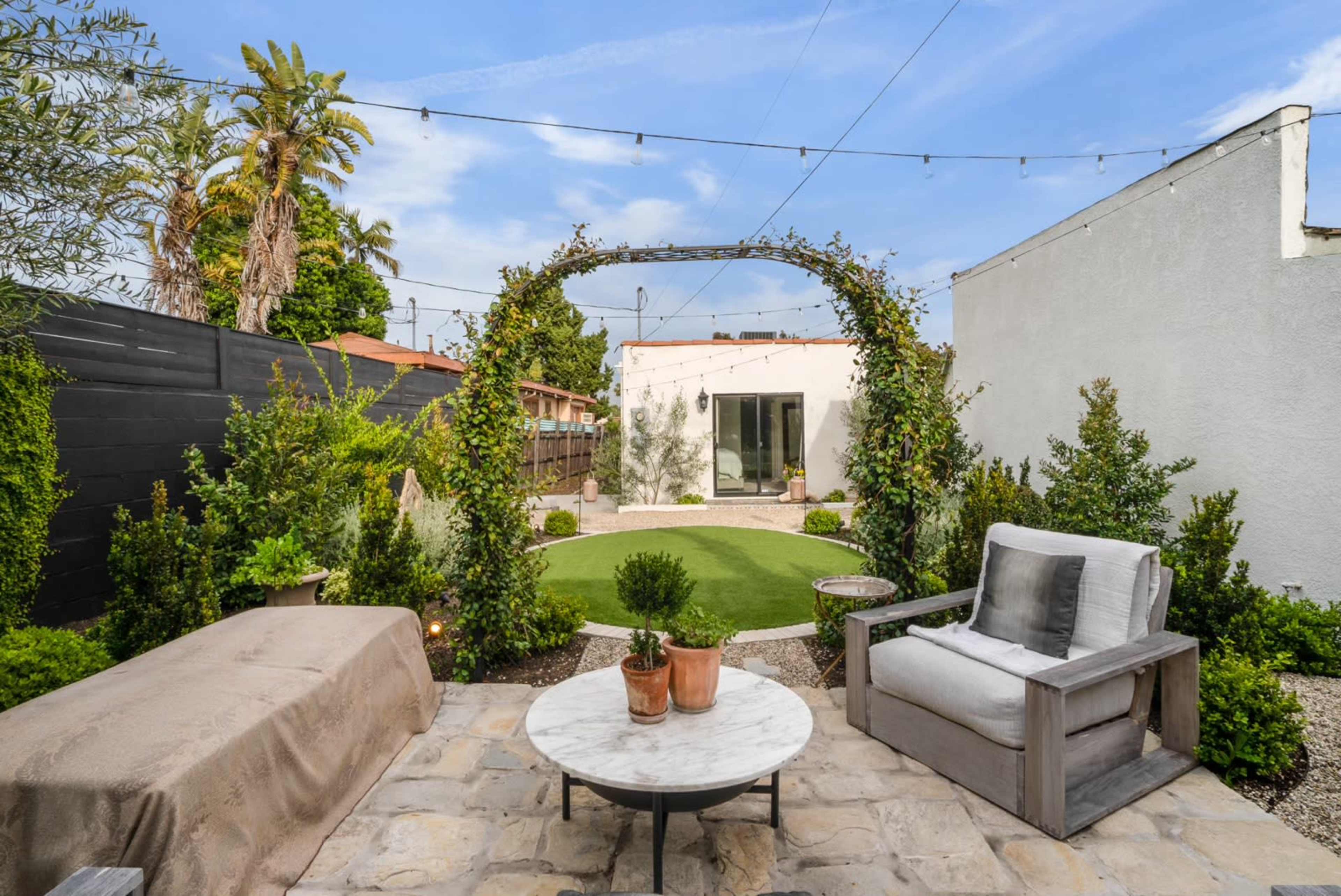 A backyard garden features a circular archway covered in greenery, surrounded by plants and a seating area with a marble table.