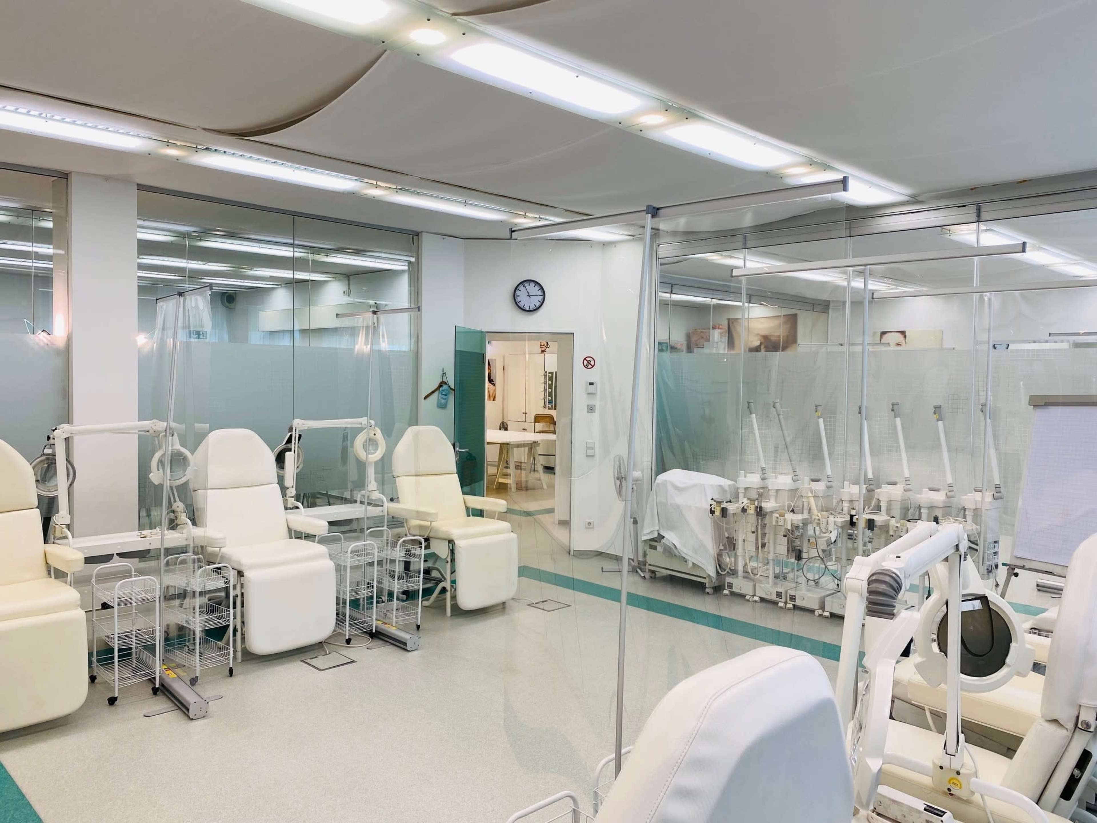 A medical room with multiple treatment stations separated by clear partitions, featuring white reclining chairs and medical equipment in the background.