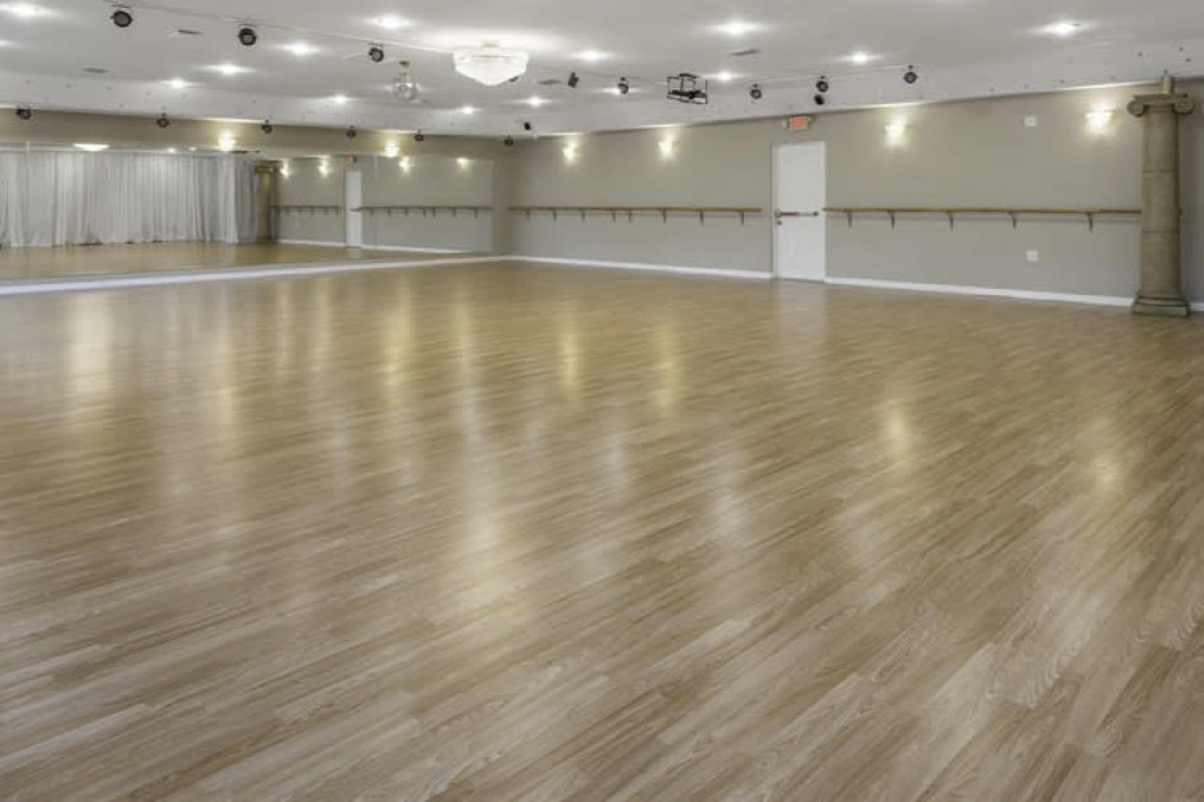 The image shows a spacious dance studio with a polished wooden floor, mirrored walls, and bright overhead lighting.