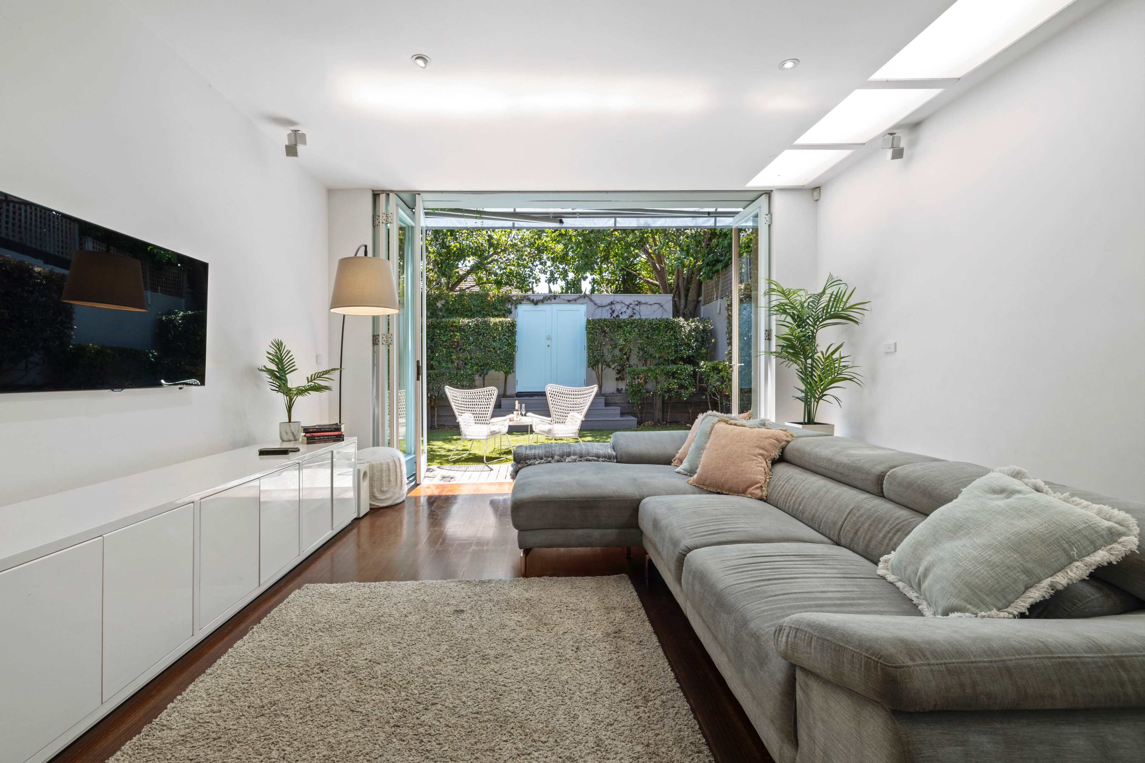 A modern living room features a large sectional sofa, a wall-mounted television, and sliding glass doors that open to a garden with two lounge chairs.