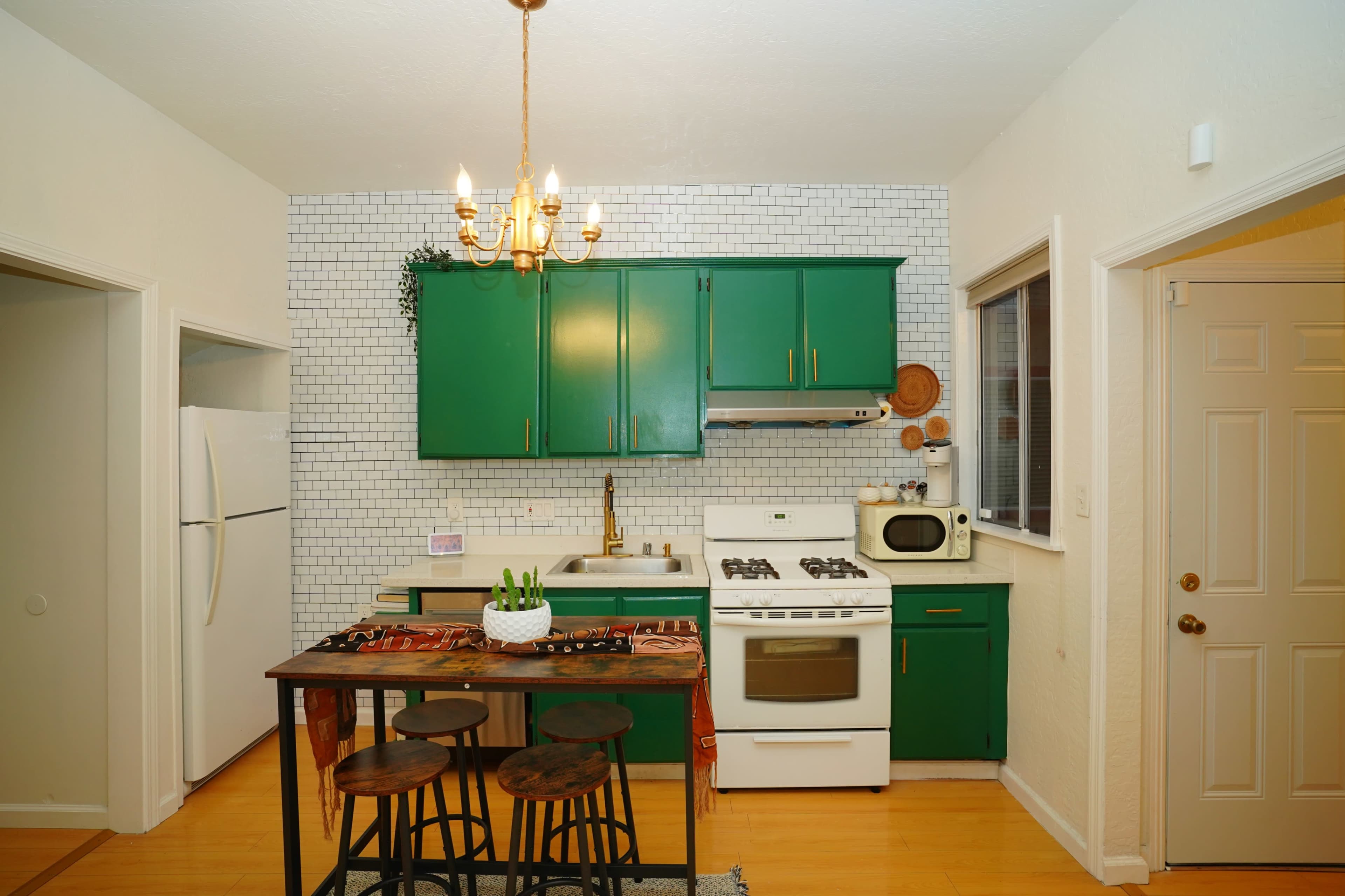 The kitchen features green cabinets, a white stove and refrigerator, a small dining table with three stools, and a tiled backsplash.