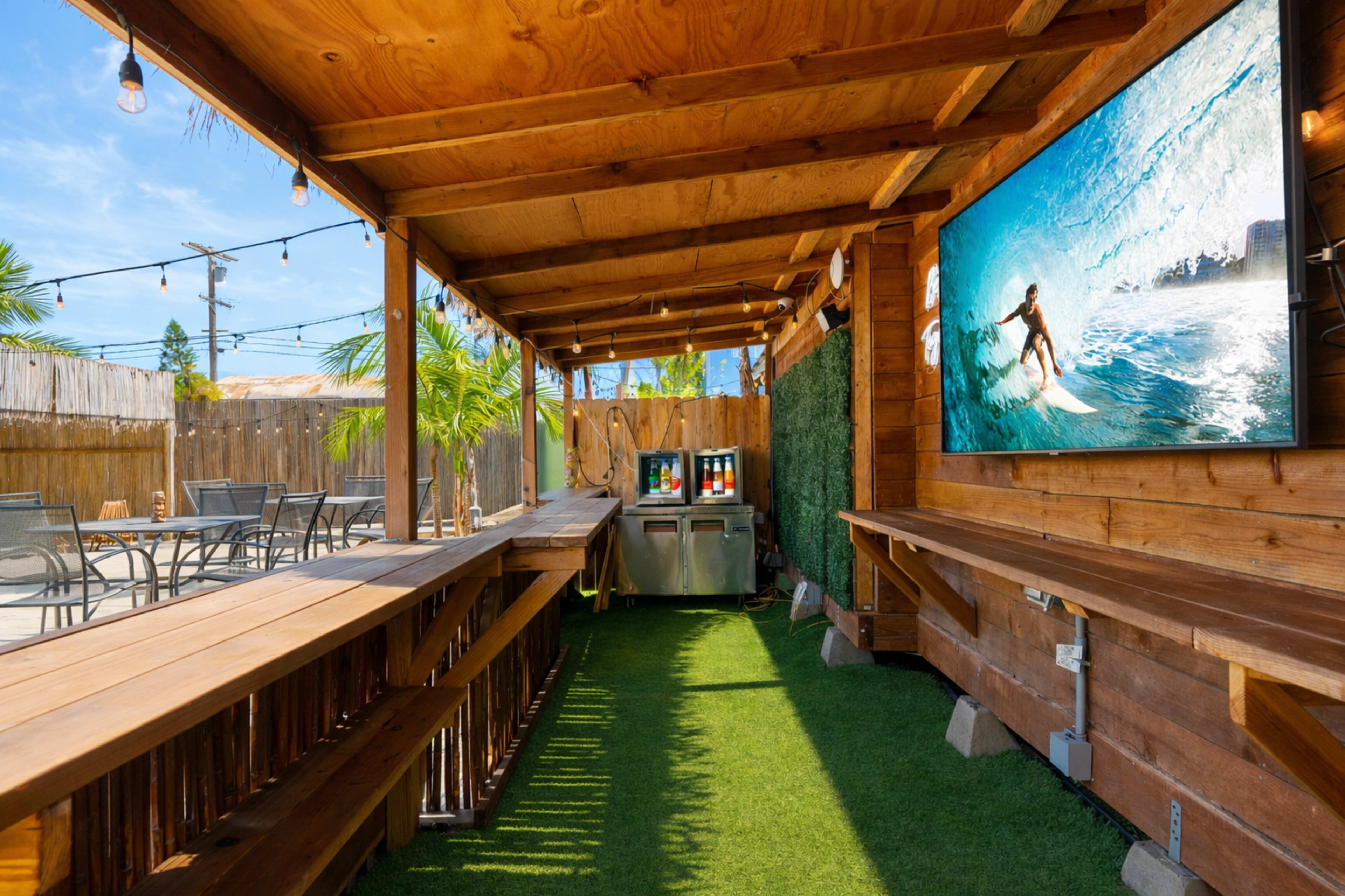 The image shows an outdoor patio area with wooden seating, a large wall-mounted TV displaying a surfing scene, and an adjacent service area with drink dispensers, all under a wooden roof.