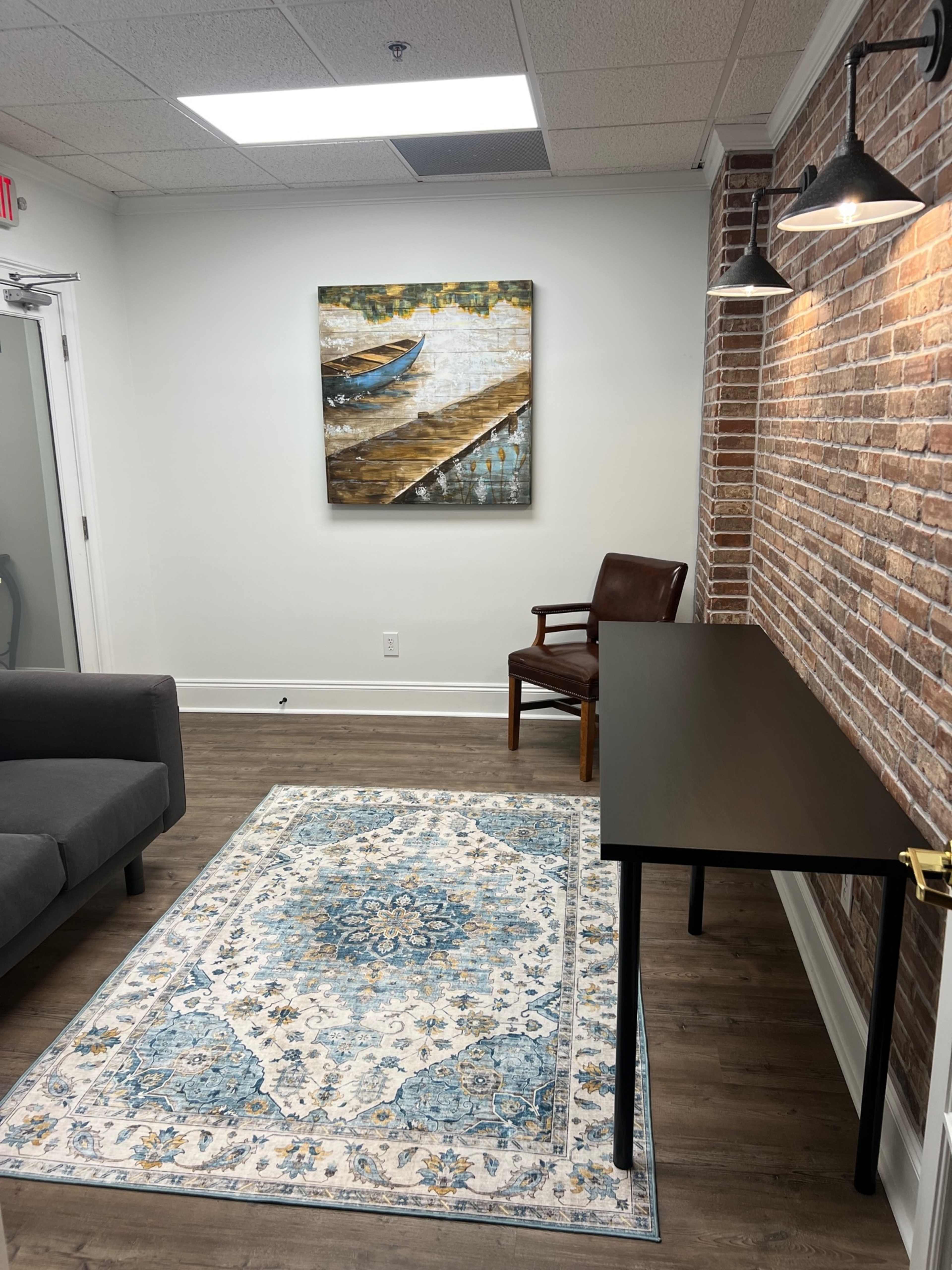 A small office space featuring a gray sofa, a wooden desk, a chair, a decorative rug, and a painting of a boat on a dock on the wall.