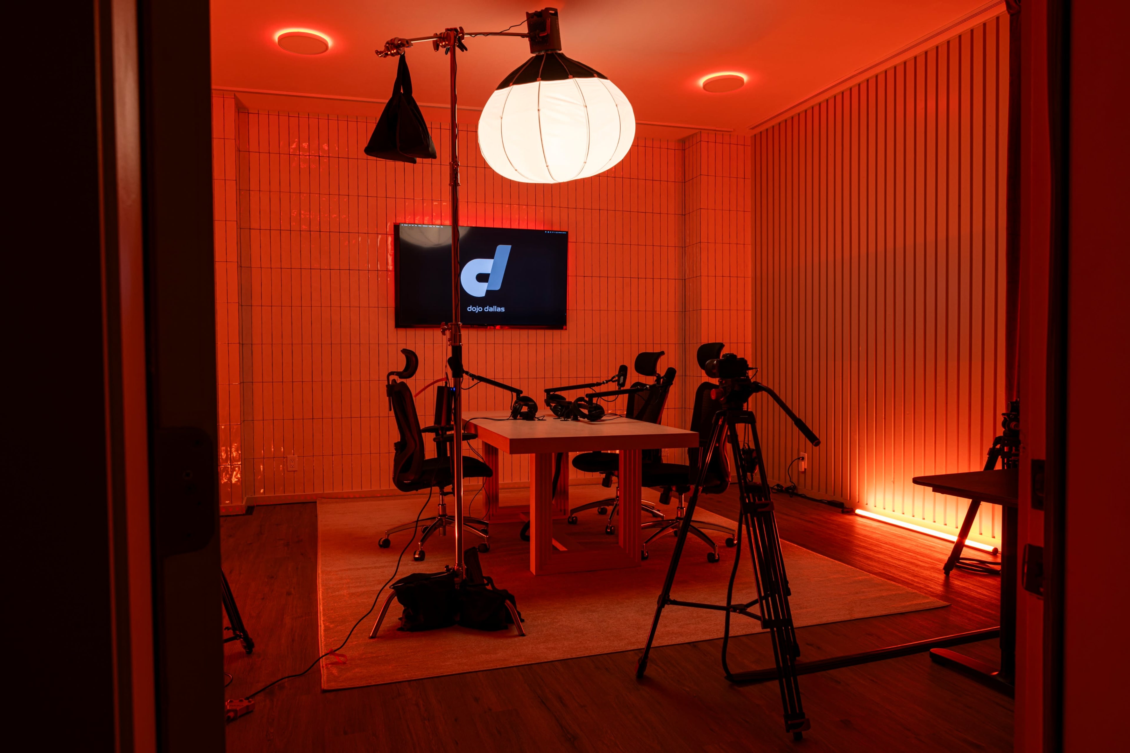 A modern recording studio with a large table, chairs, multiple cameras, and a bright circular light, all set against an orange backdrop.