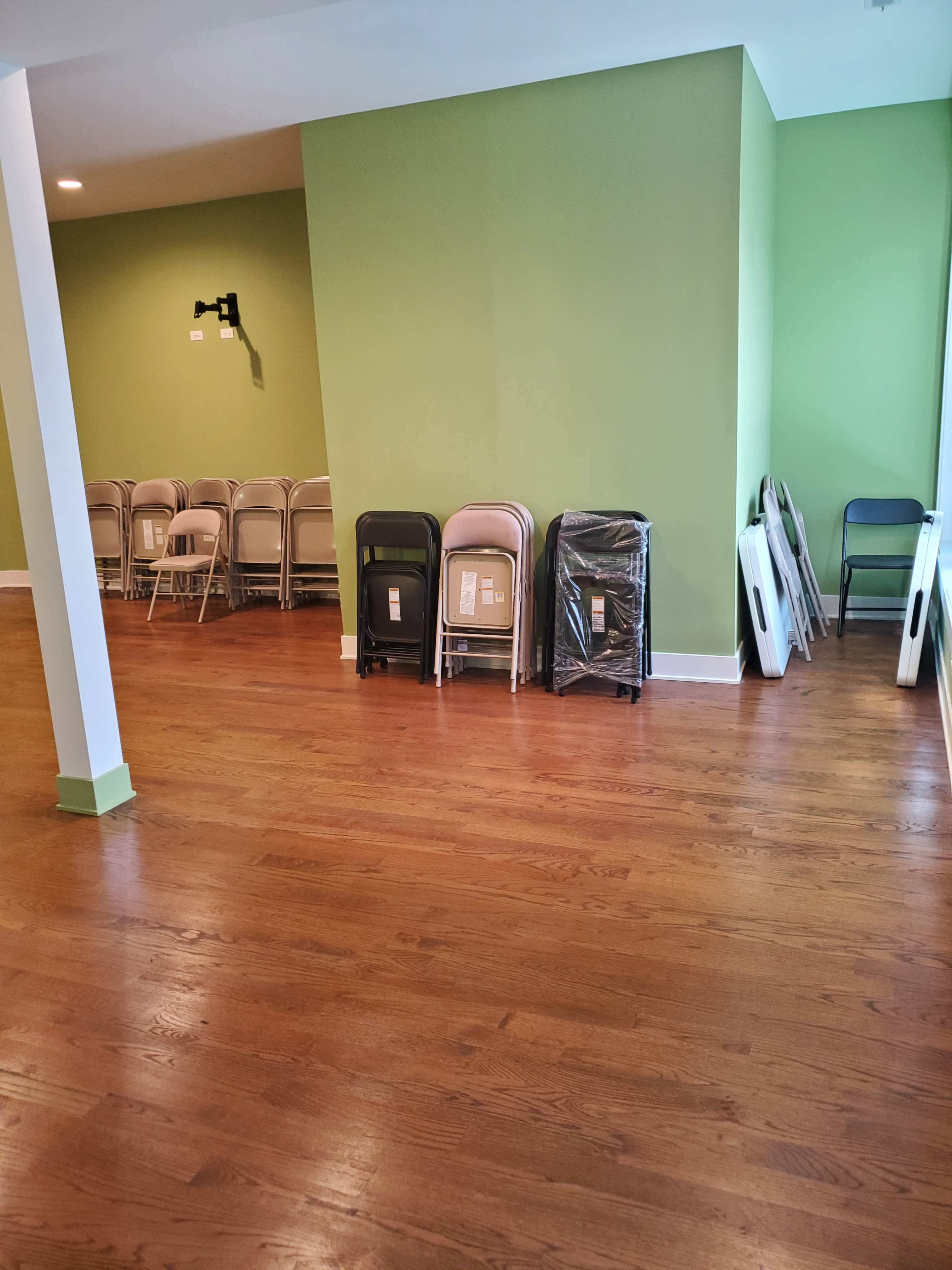 The image shows a spacious room with wooden flooring, featuring a green wall, stacked folding chairs, and some unoccupied tables along one side.
