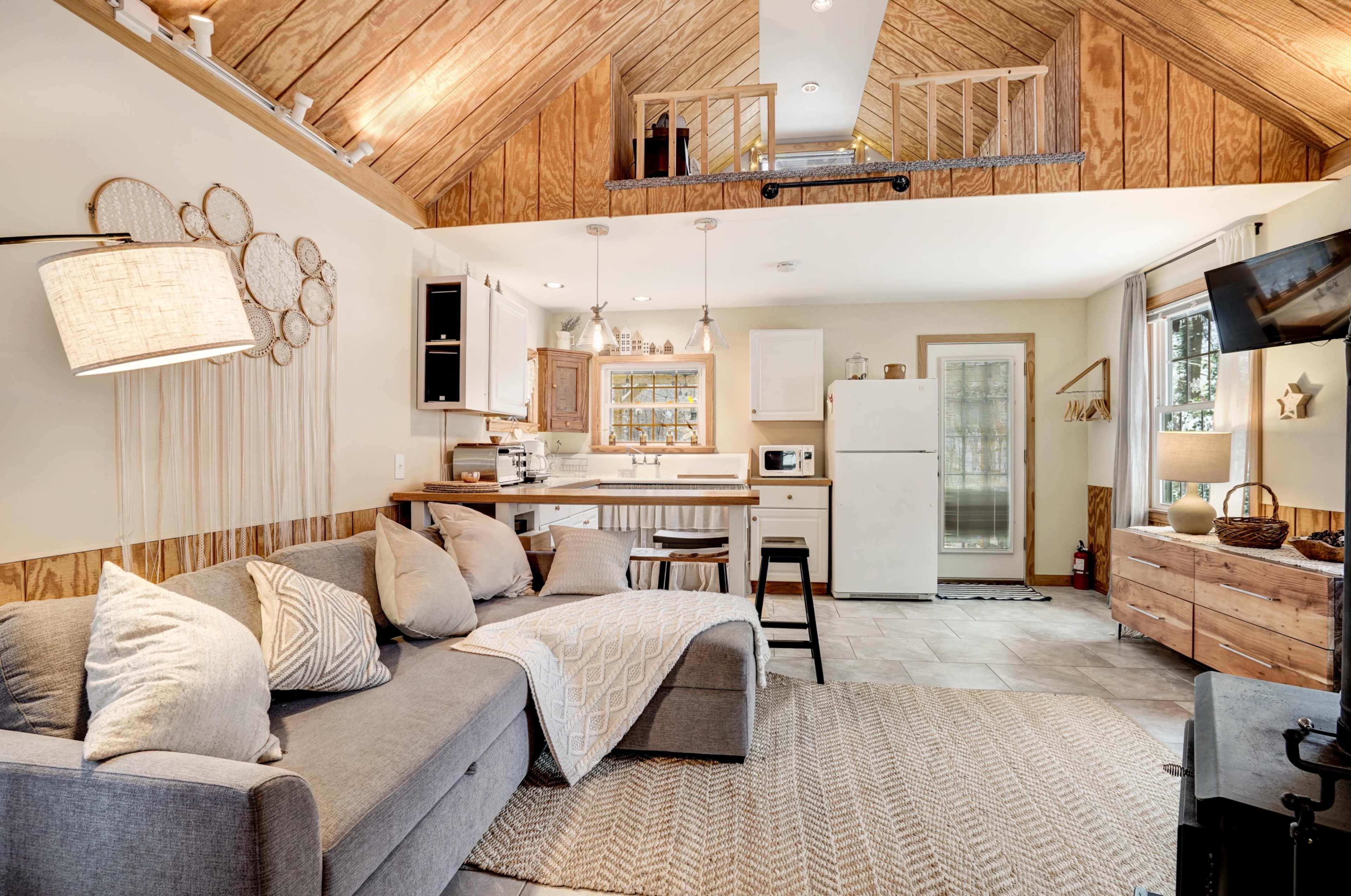 A cozy living space with a gray sofa, a kitchen area including a refrigerator and microwave, and wooden accents highlighted by a lofted ceiling with exposed beams.