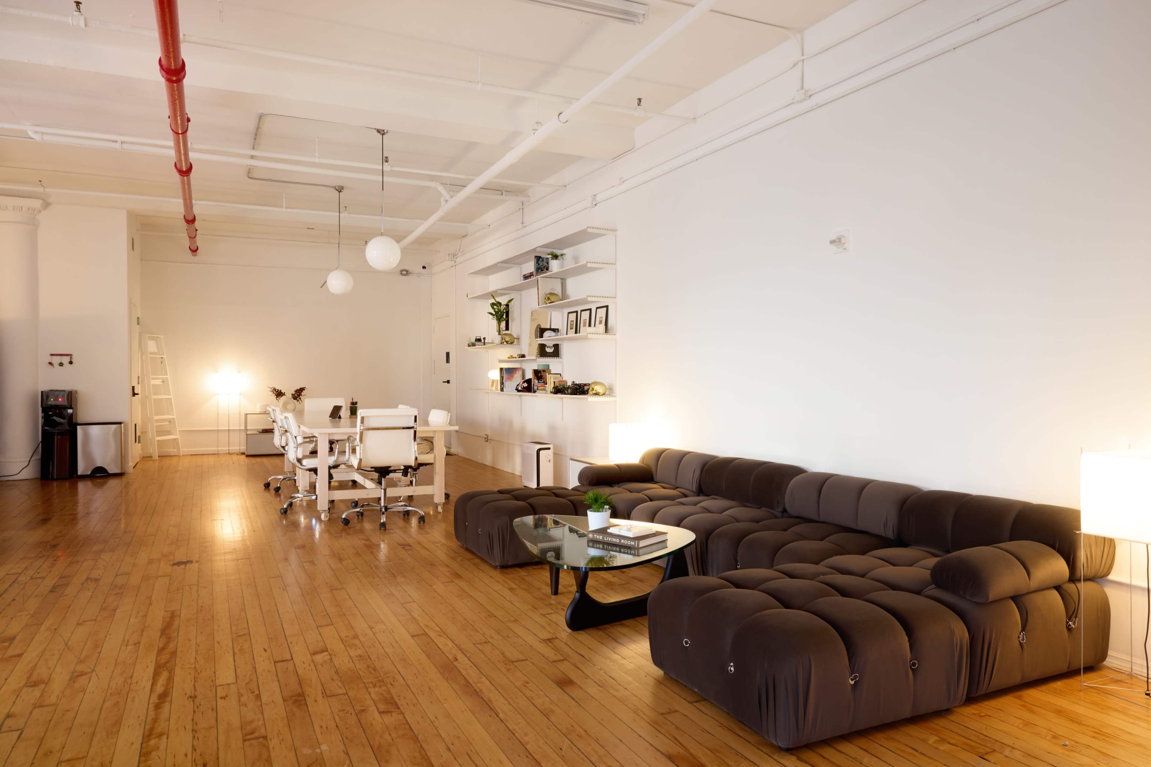 The image shows a spacious, minimalist office interior featuring a brown sectional sofa, a glass coffee table, and a workspace with white desks and chairs.