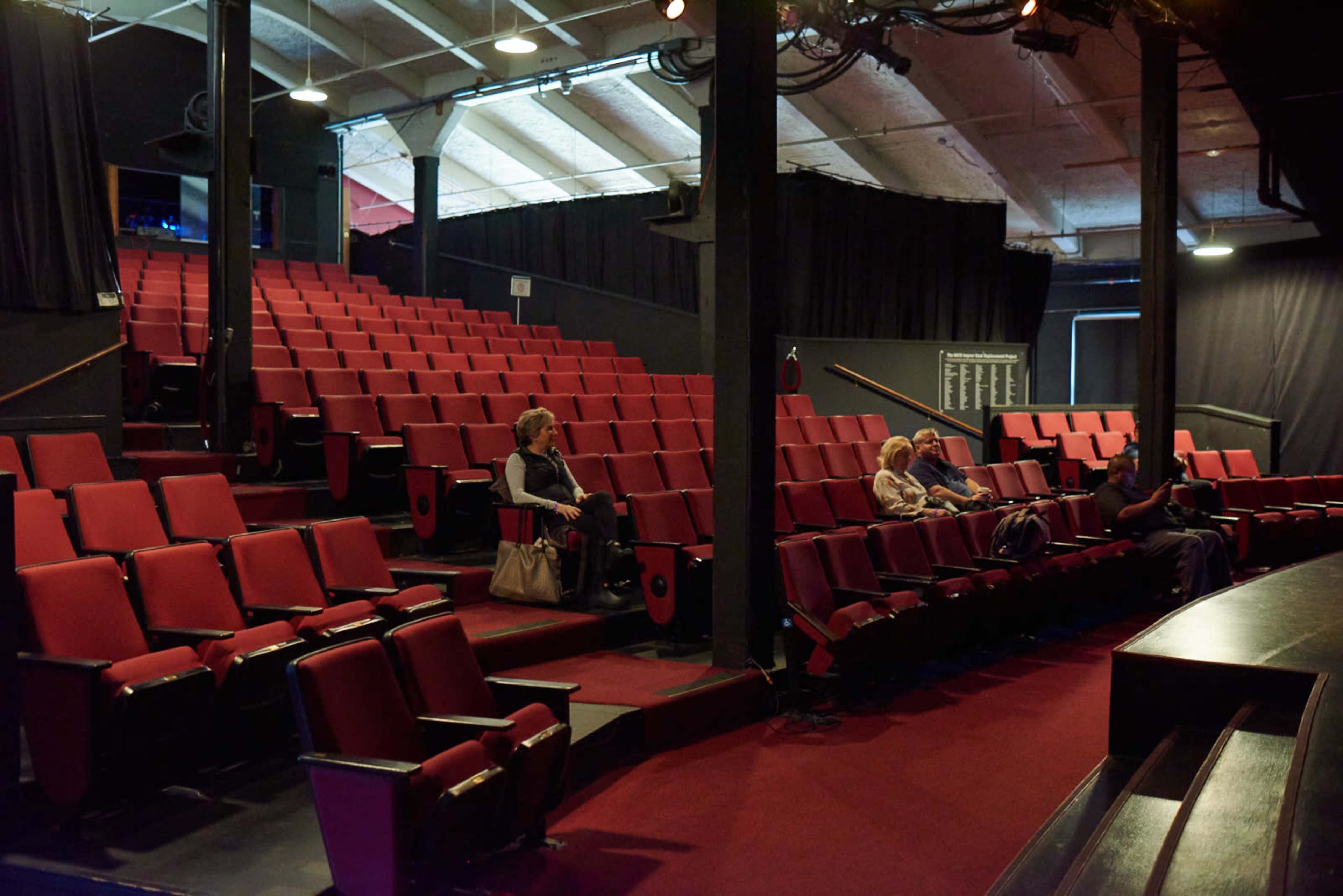 An empty theater with red seats and a sparse audience seated in the front rows.