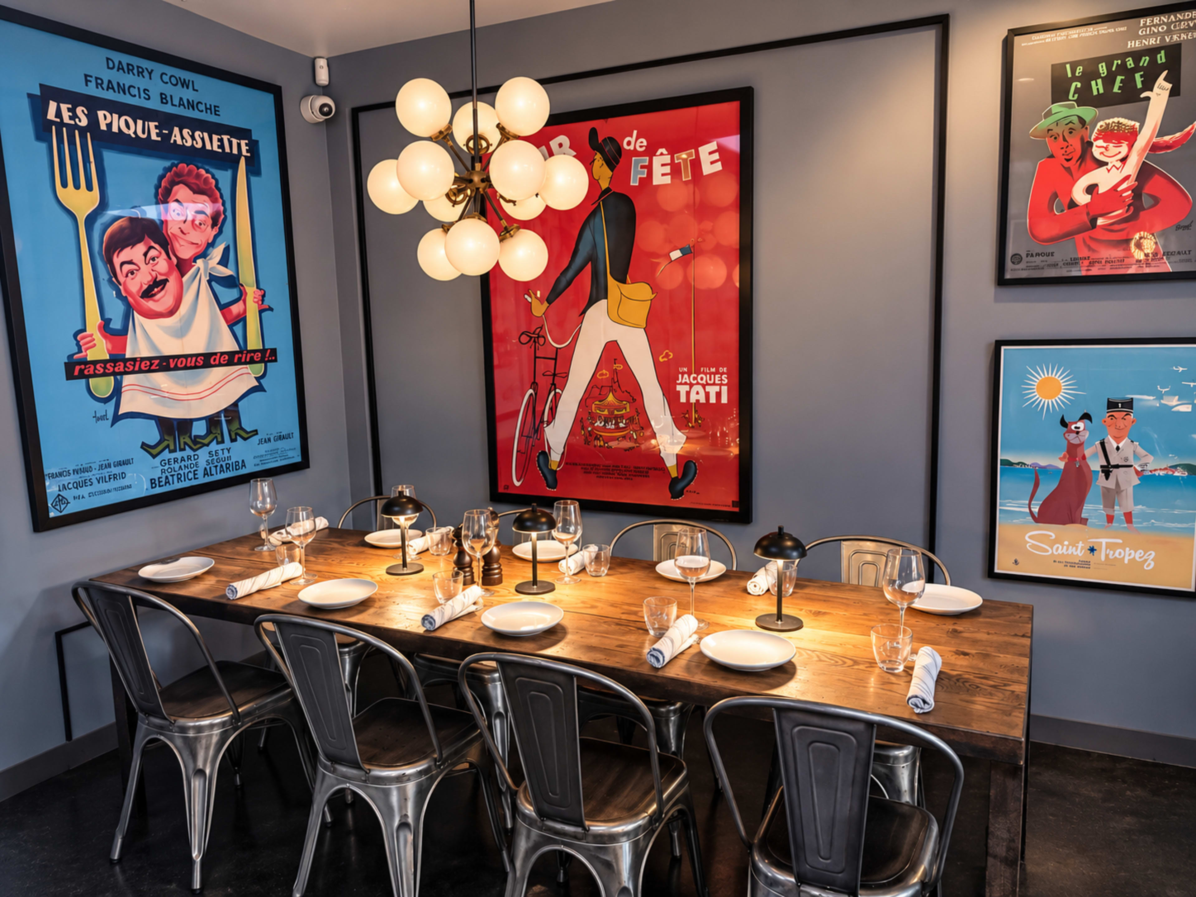 A dining area features a long wooden table set for dinner, surrounded by metal chairs and adorned with vintage posters on the walls and a modern light fixture above.