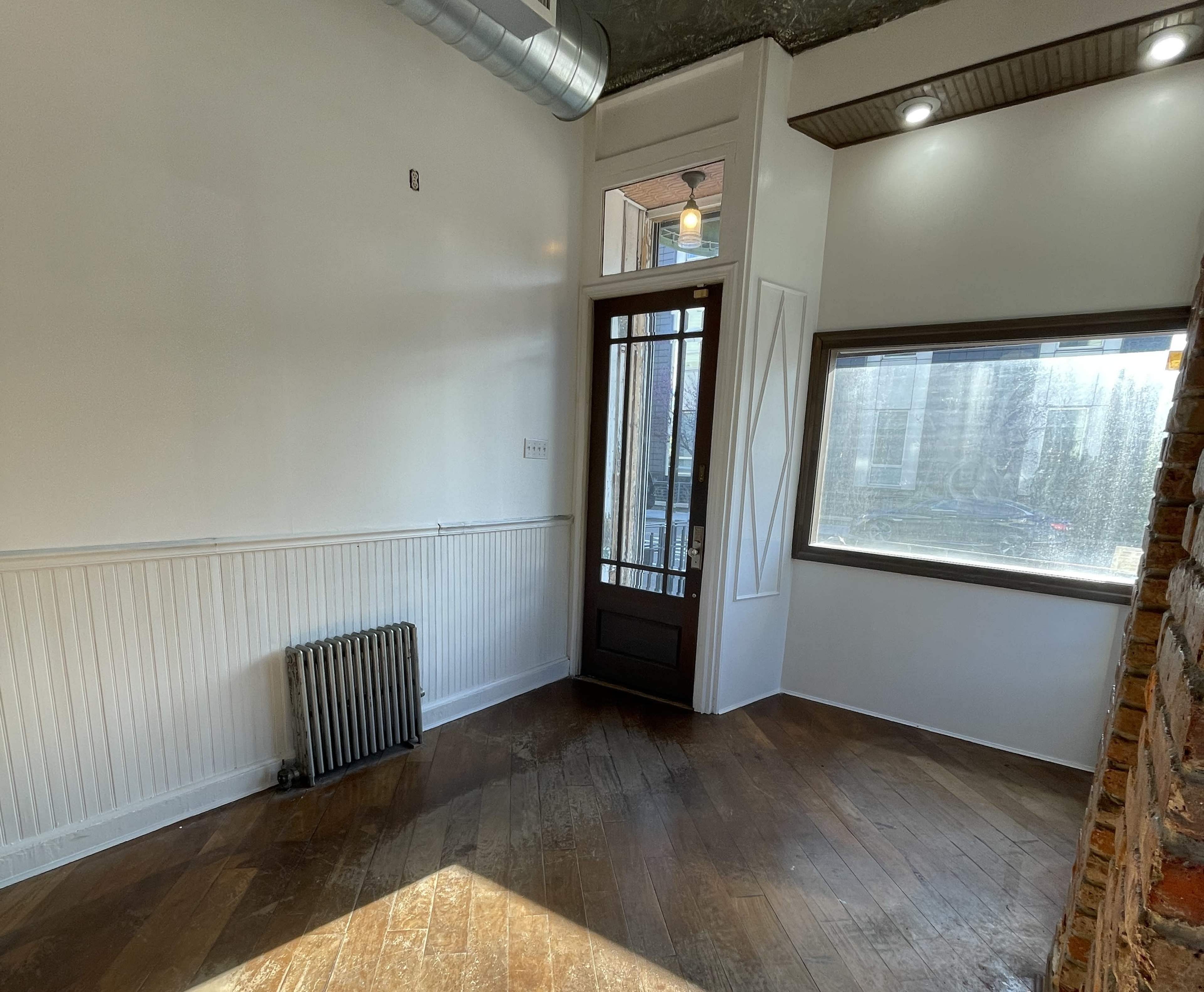 The image shows a small room with wooden flooring, a large window, a door with glass panels, and a radiator against one wall.
