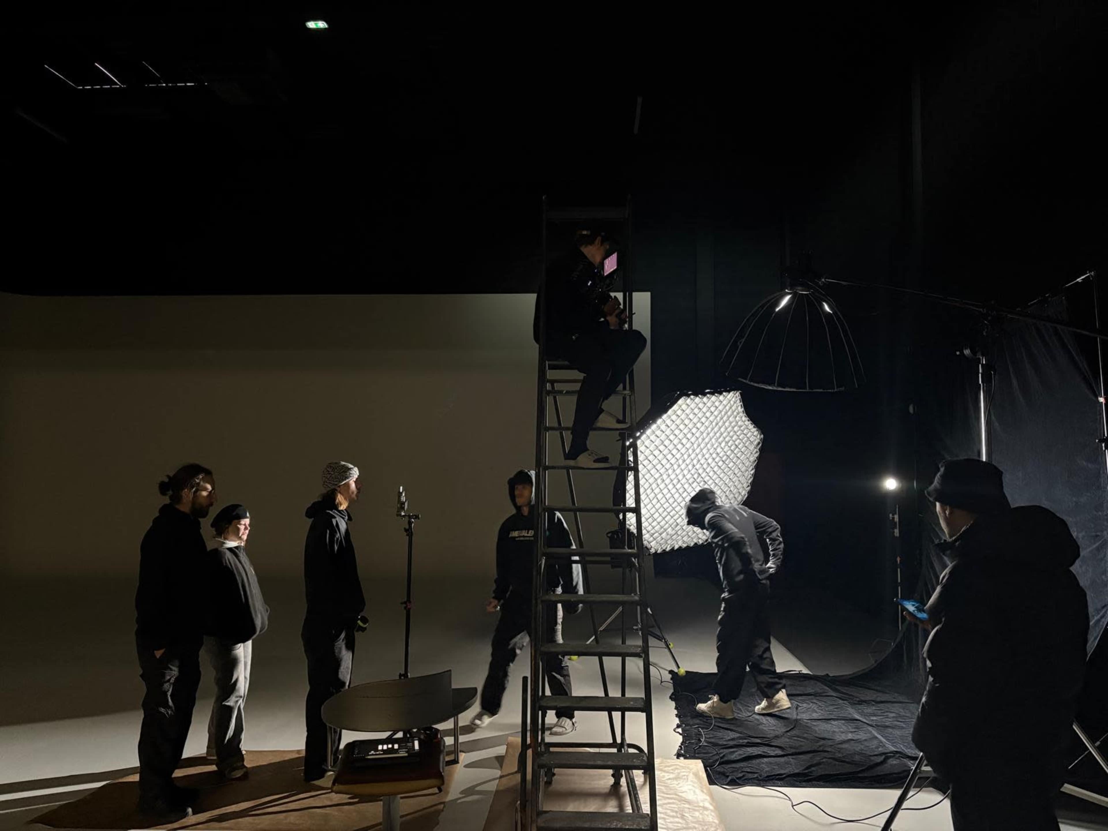 A group of people is gathered in a studio, with one person on a ladder adjusting equipment while others are positioned nearby, and a large light source is set up to the side.