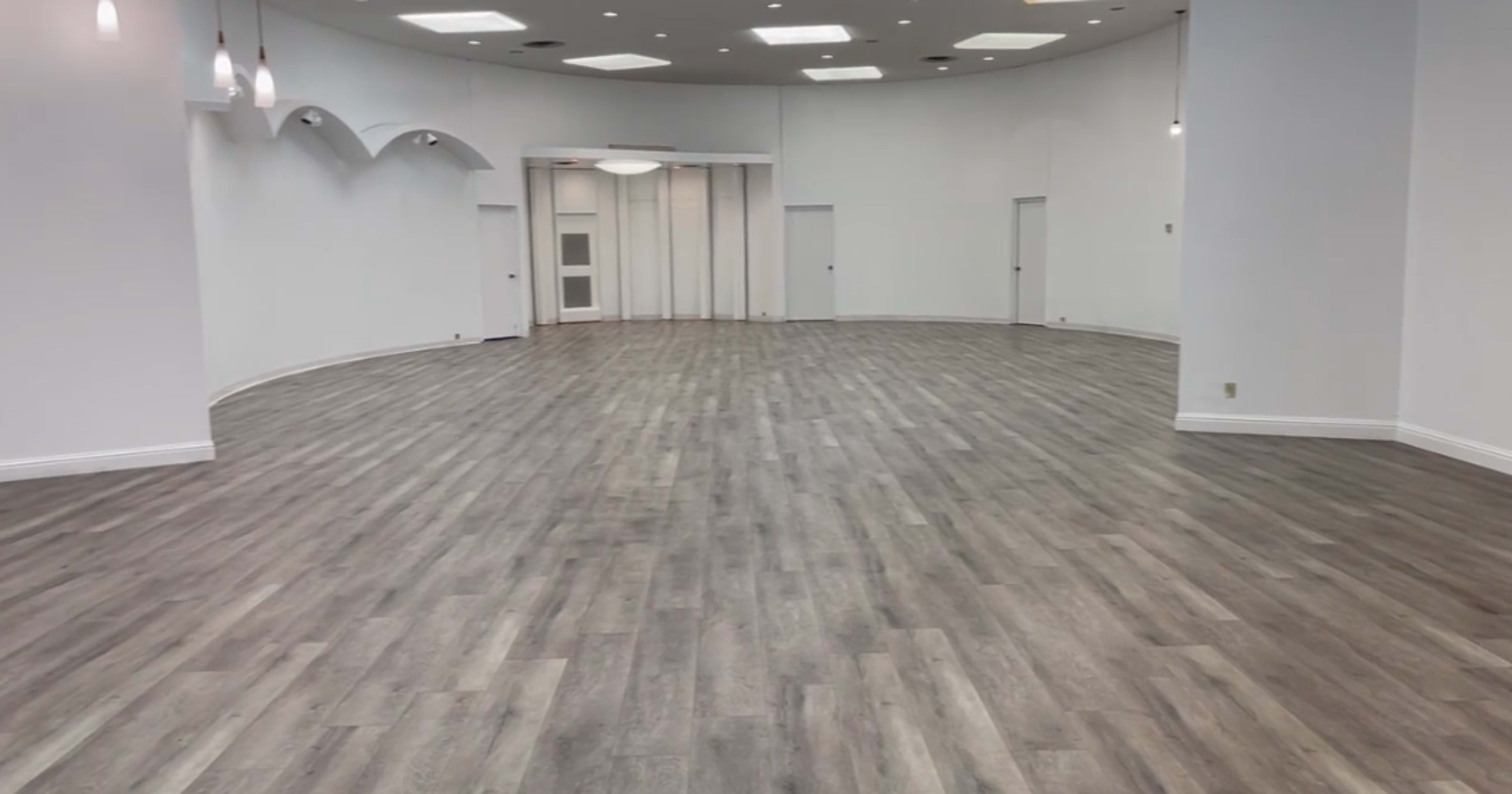A spacious, empty room with light-colored wooden flooring and white walls, featuring multiple doorways and overhead lighting.