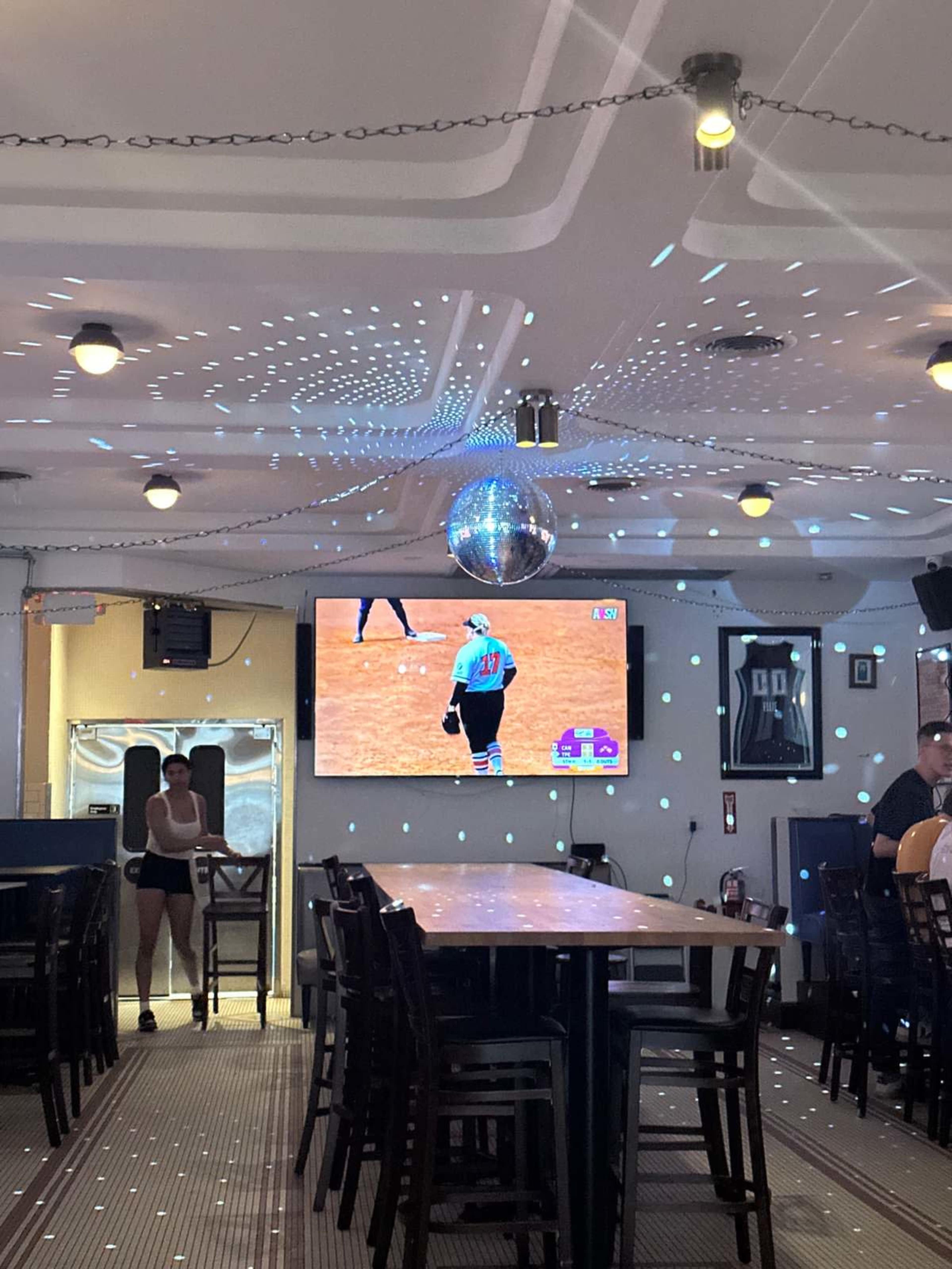 The image shows a restaurant or bar interior with a large screen displaying a sports game, a disco ball hanging from the ceiling, and patrons seated at a long table.