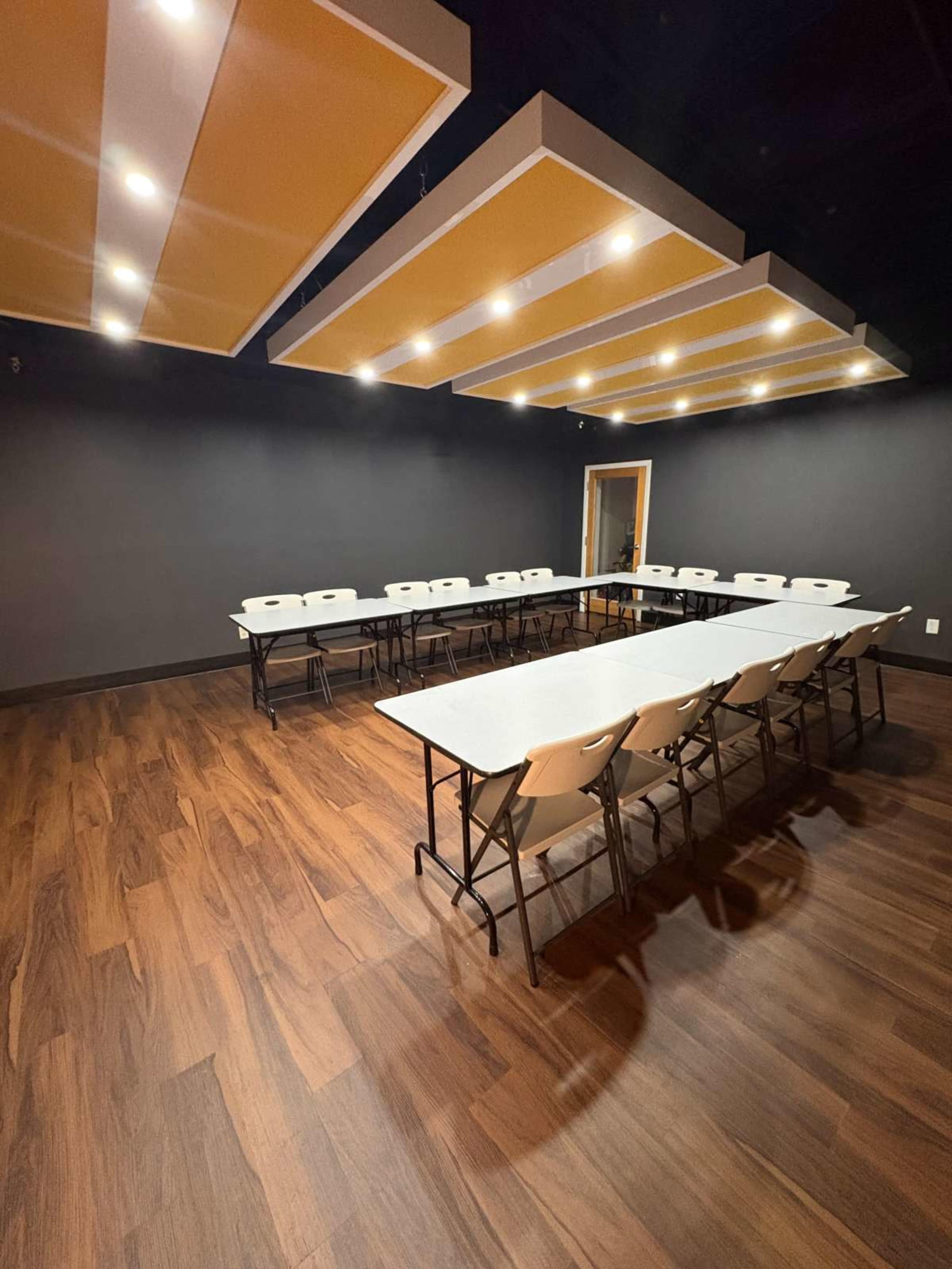 The image shows a conference room set up with several folding tables arranged in a U-shape, surrounded by white chairs, under bright ceiling lights.