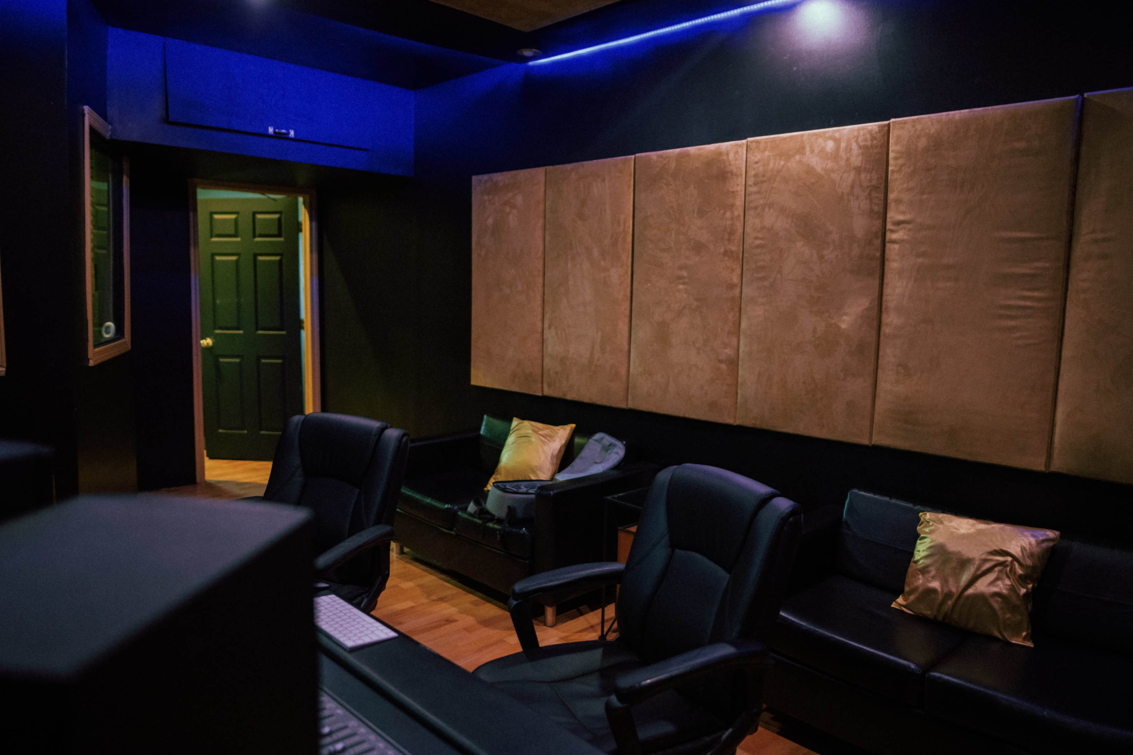 A dimly lit recording studio with black walls, two leather chairs with decorative pillows, and a green door at the far end.