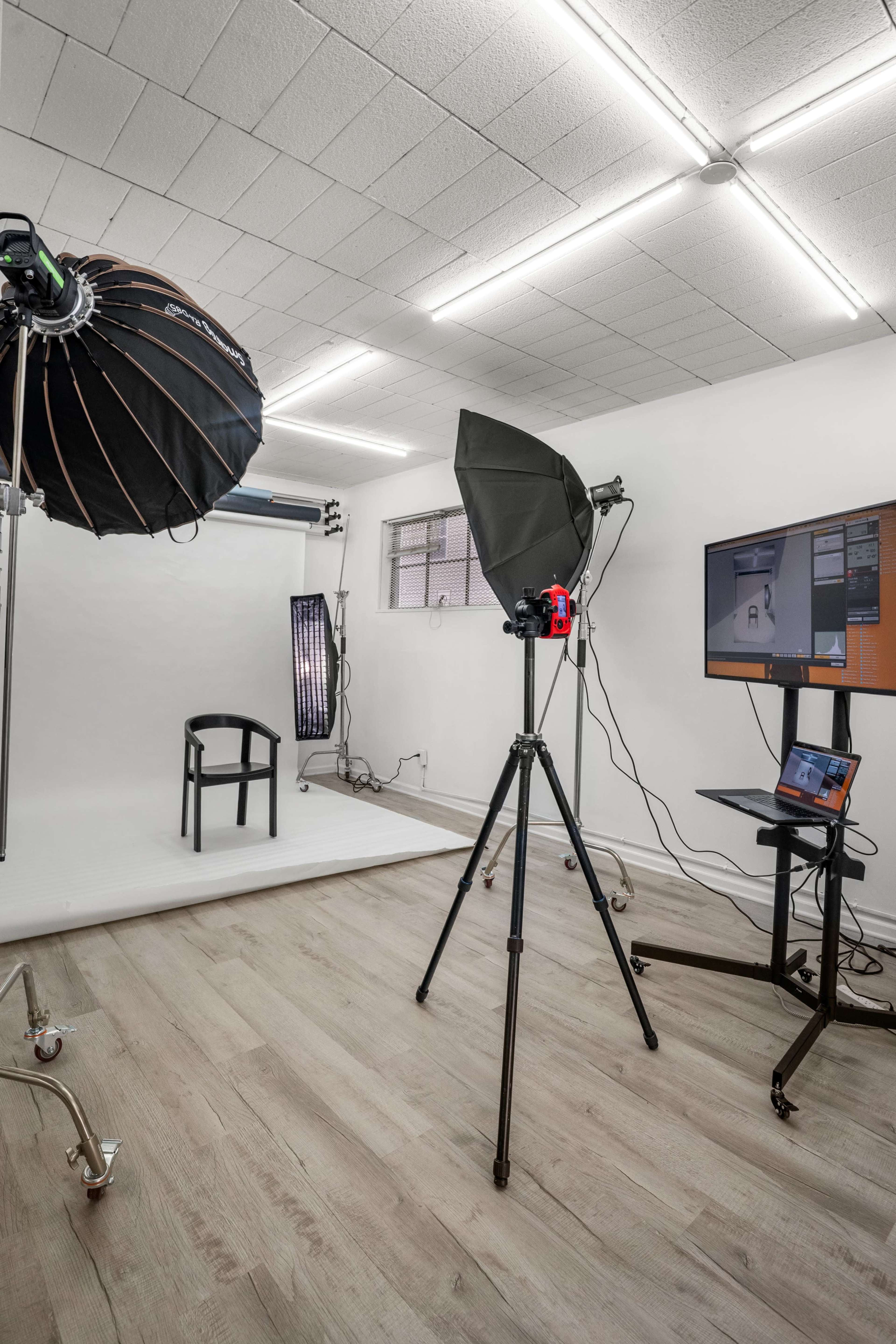 The image shows a photography studio equipped with lights, a backdrop, a chair, and a computer on a stand.