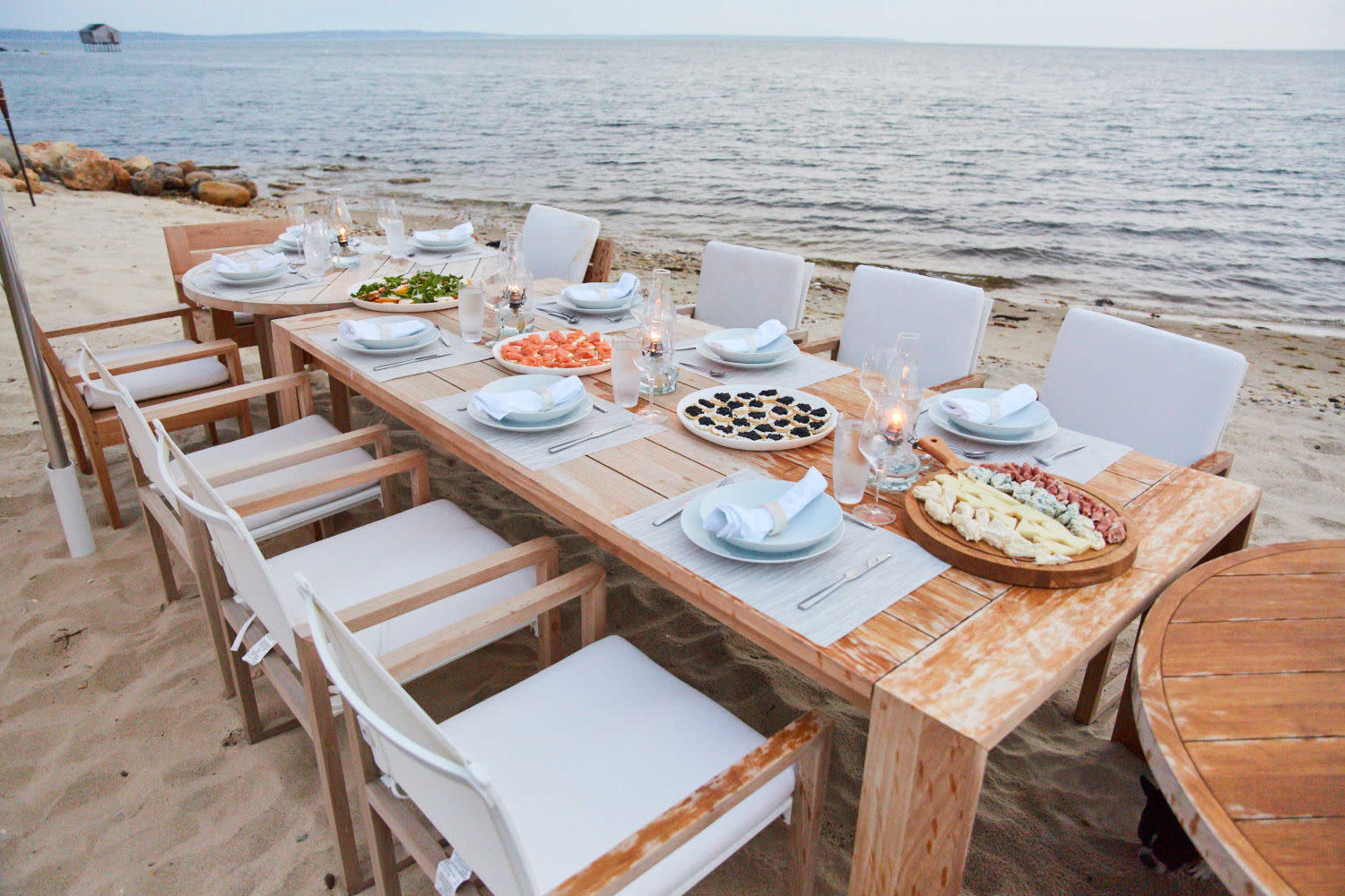 A long wooden dining table is set for a meal on the beach, surrounded by white chairs, with various dishes and candles arranged on top.