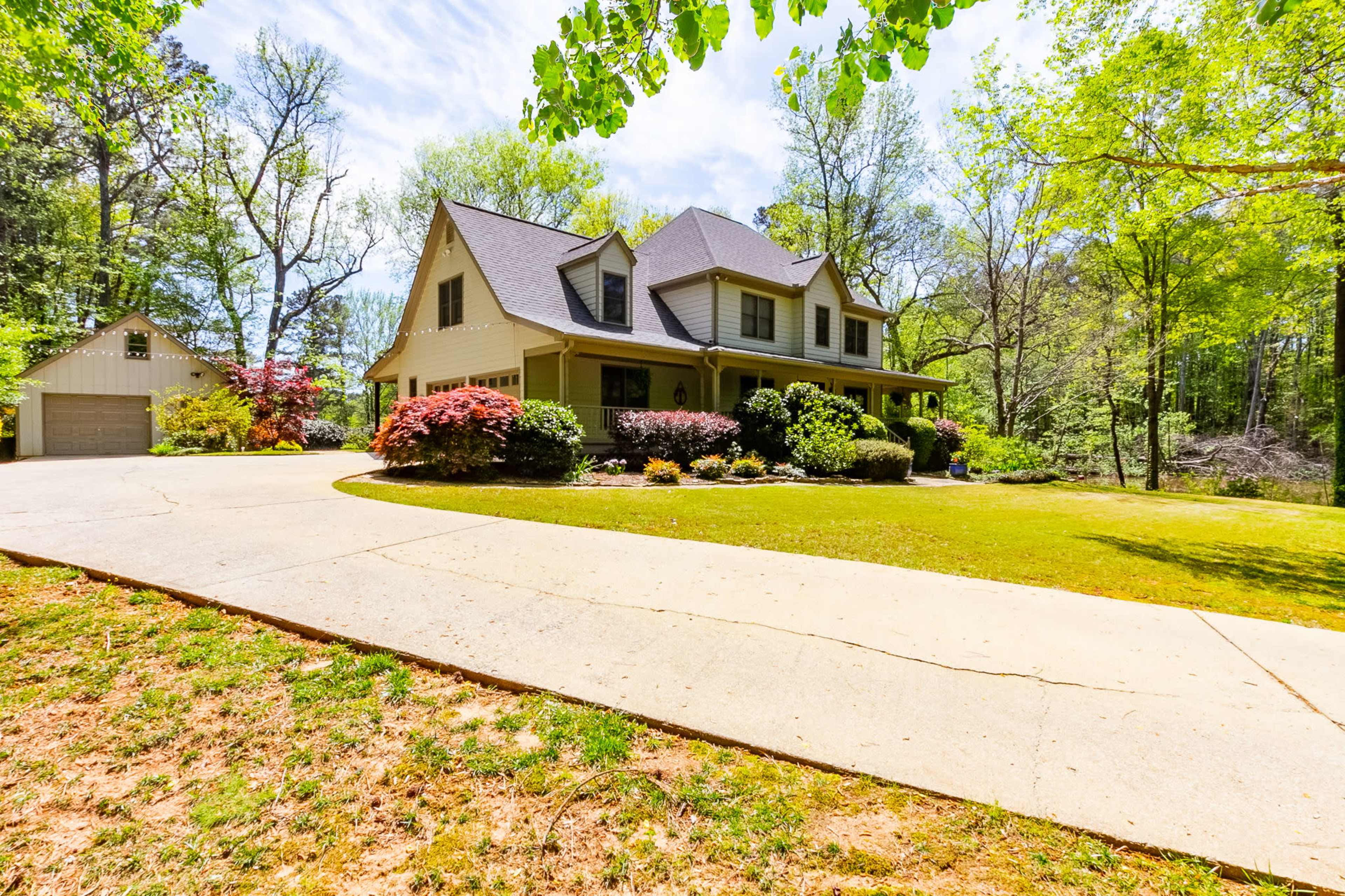 A landscaped driveway leads to a two-story house surrounded by trees and flowering shrubs.