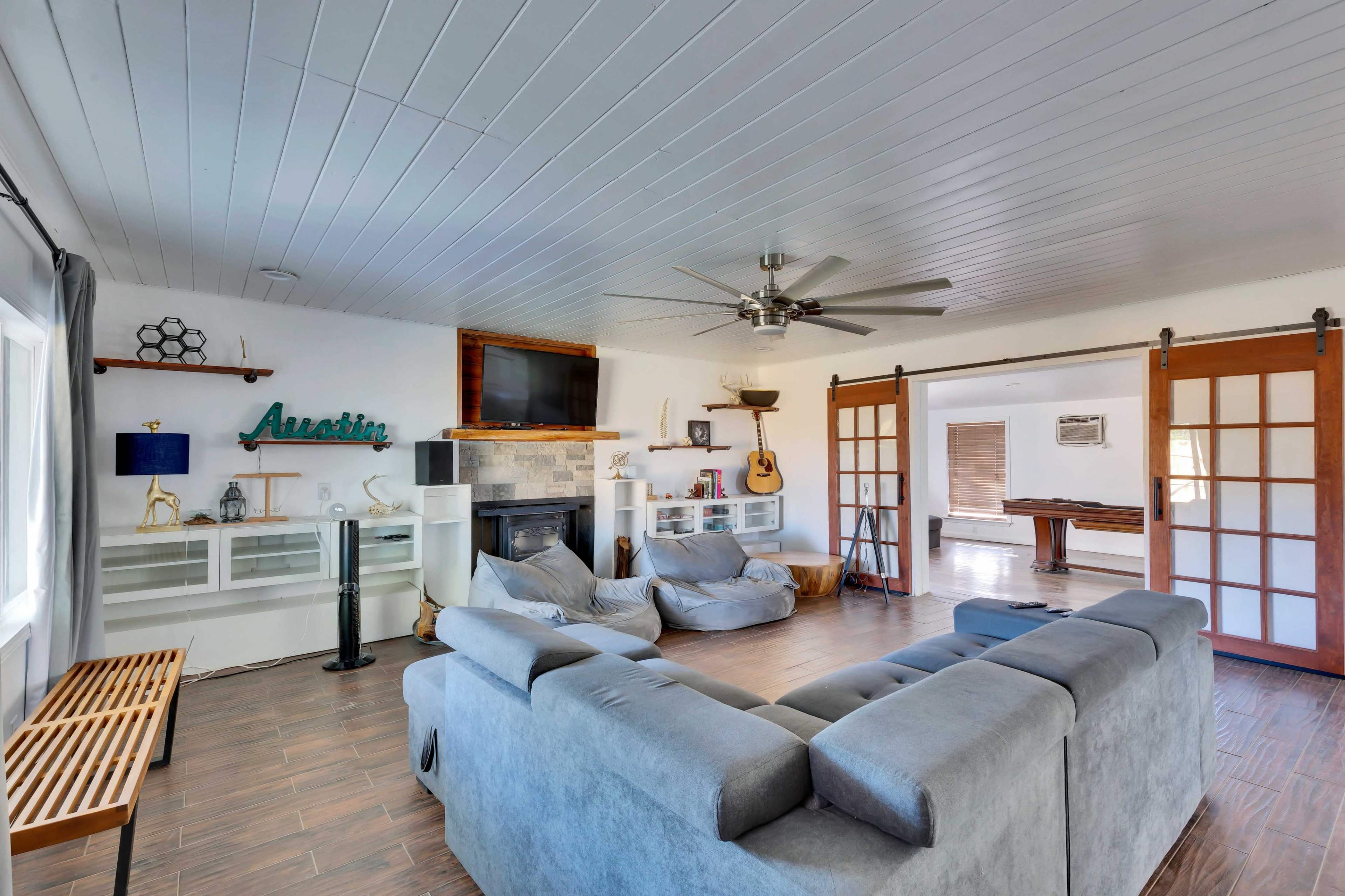 A spacious living room features a ceiling fan, a gray sectional sofa, and a fireplace, with large windows and a set of sliding doors leading to another area.