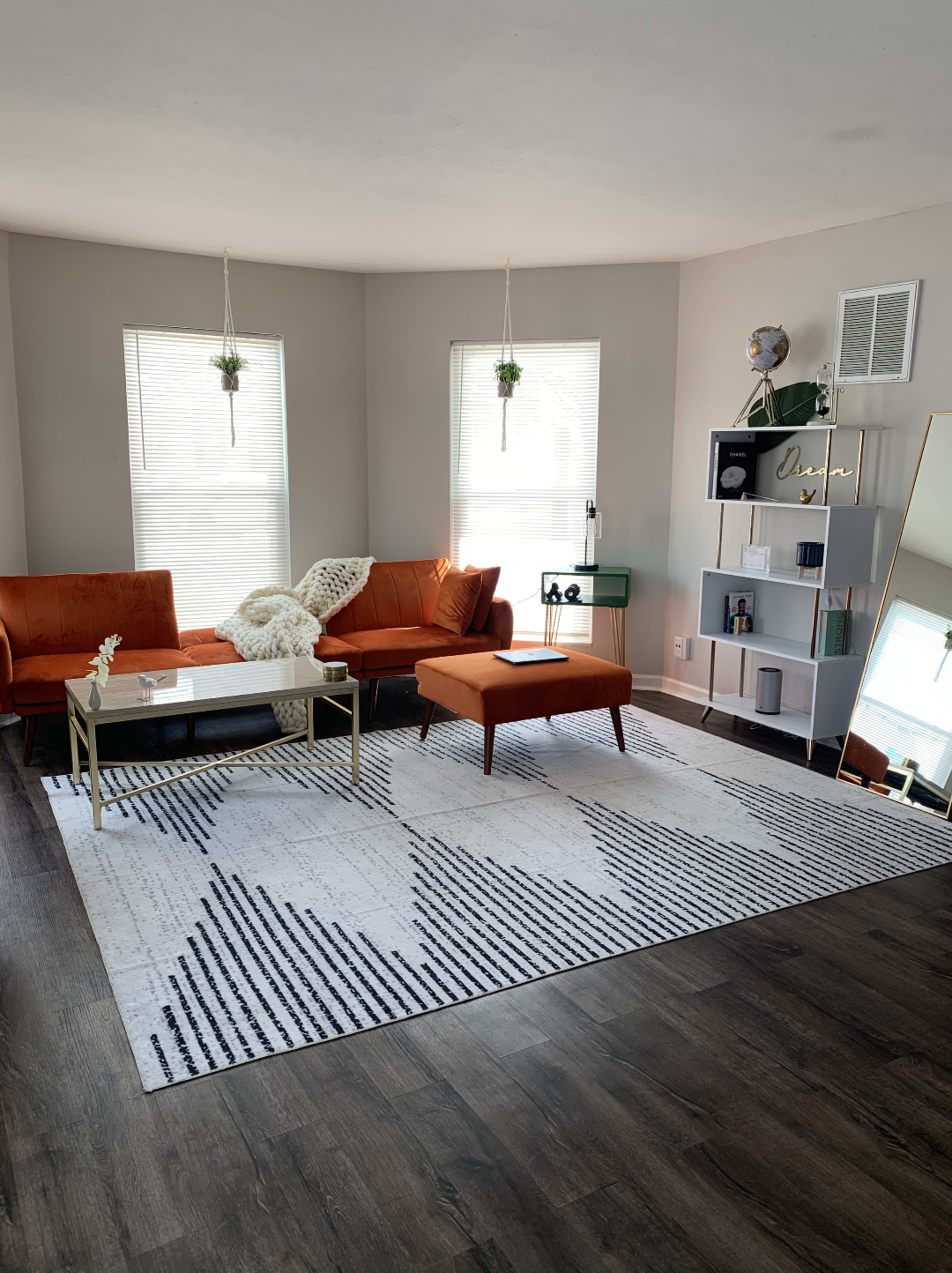The living room features a pair of orange sofas, a coffee table, a patterned rug, and a bookshelf against the wall, illuminated by natural light from two windows.