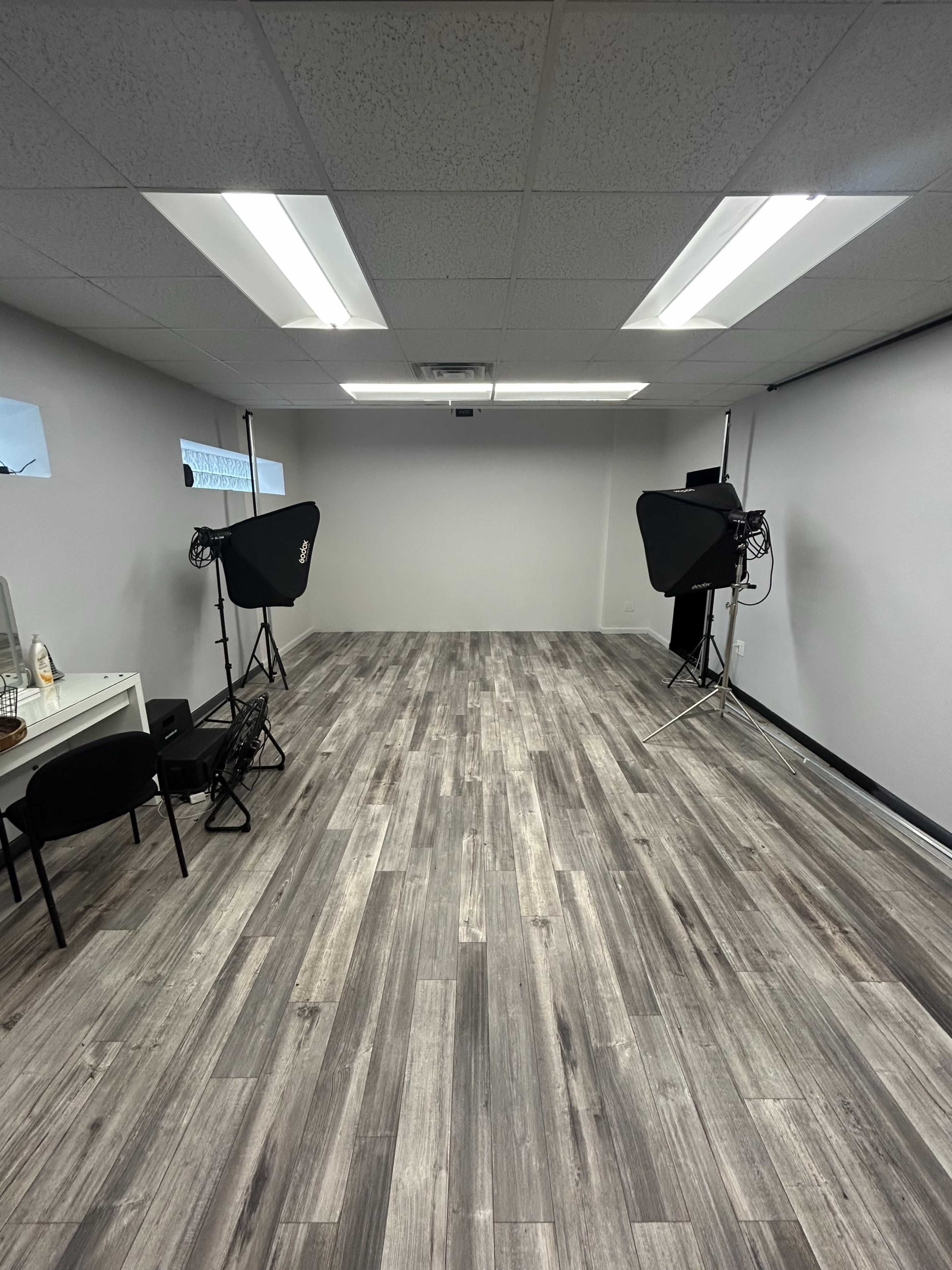 The image shows a spacious, empty room with gray walls, wooden flooring, and two photography light setups positioned on either side.