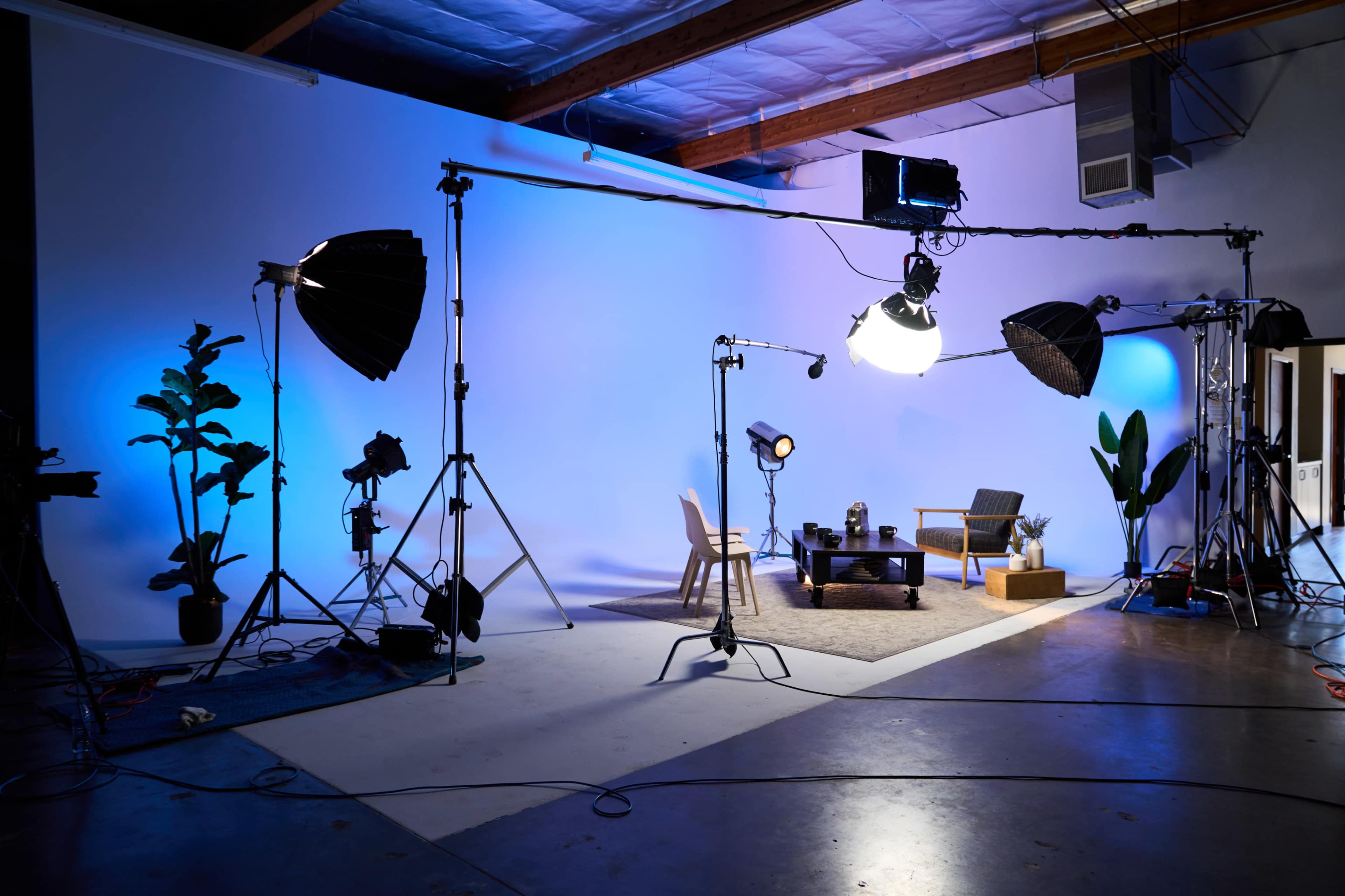 The image shows a photography studio set up with various lighting equipment, a chair, a table, and decorative plants against a light blue backdrop.