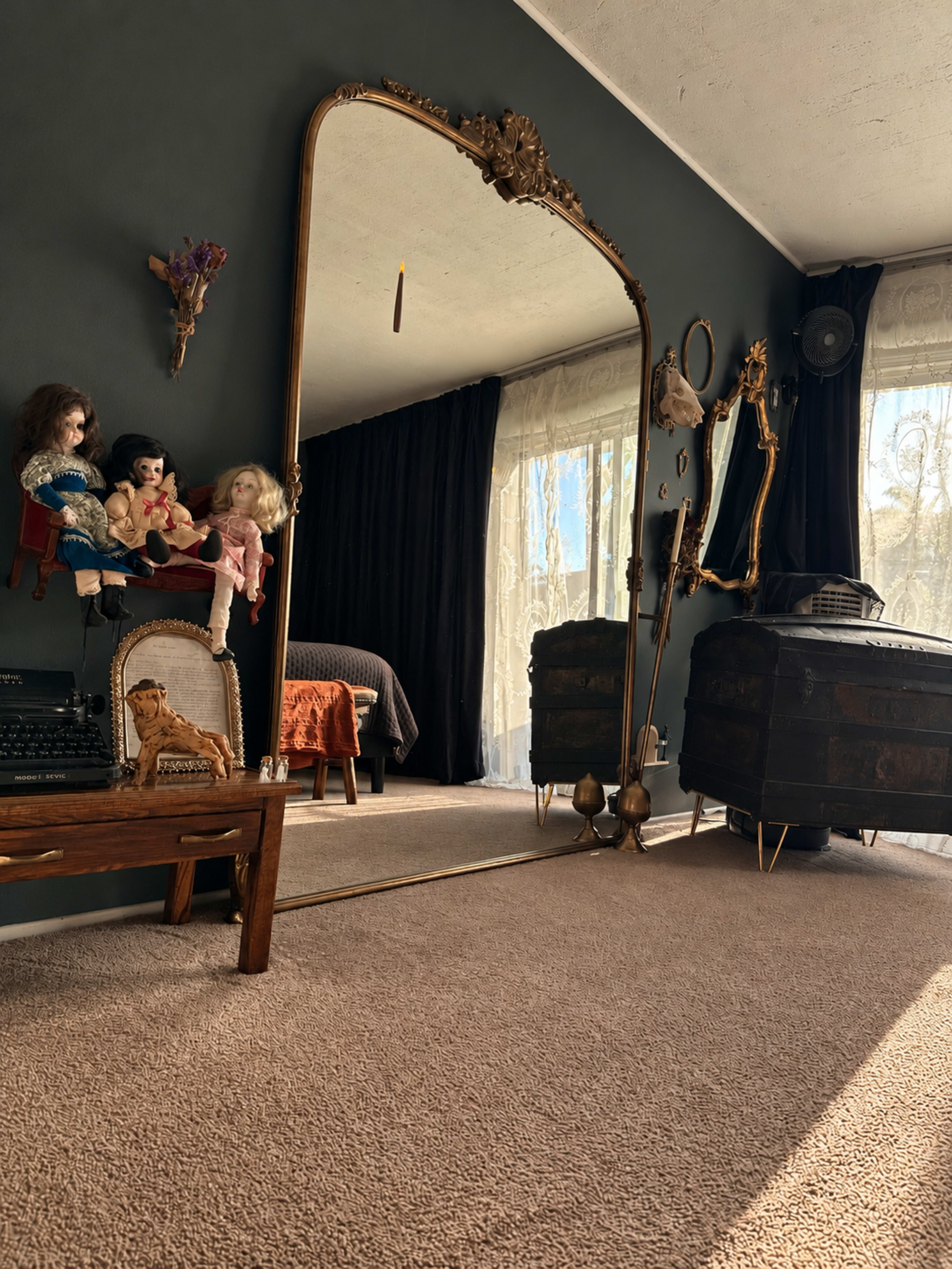 The image shows a room with a large ornate mirror reflecting a cozy space featuring a bed, curtains, and vintage dolls placed on a piece of furniture.