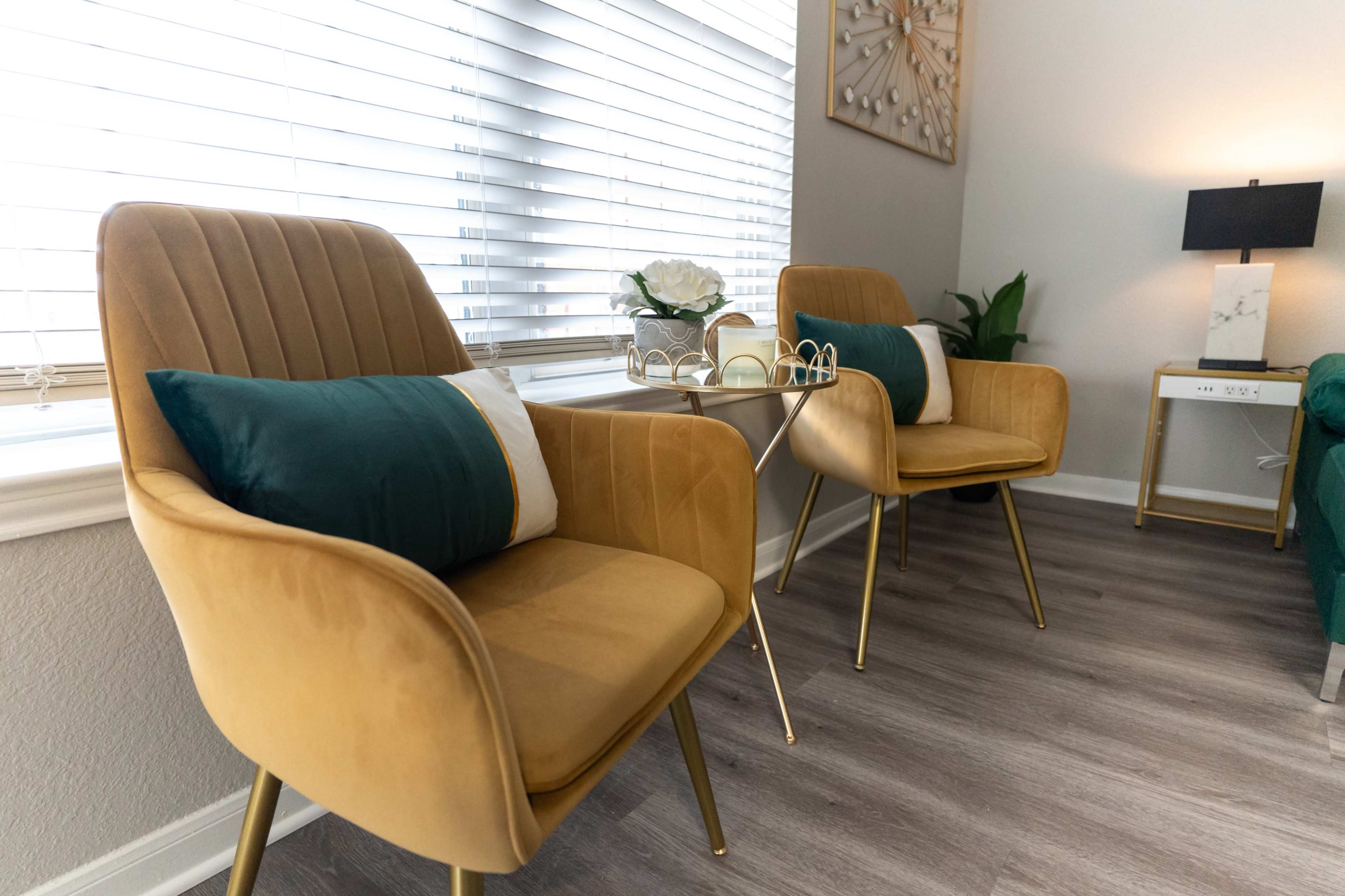 The image shows a cozy seating area with two mustard-colored chairs and a small table positioned by a window with blinds.