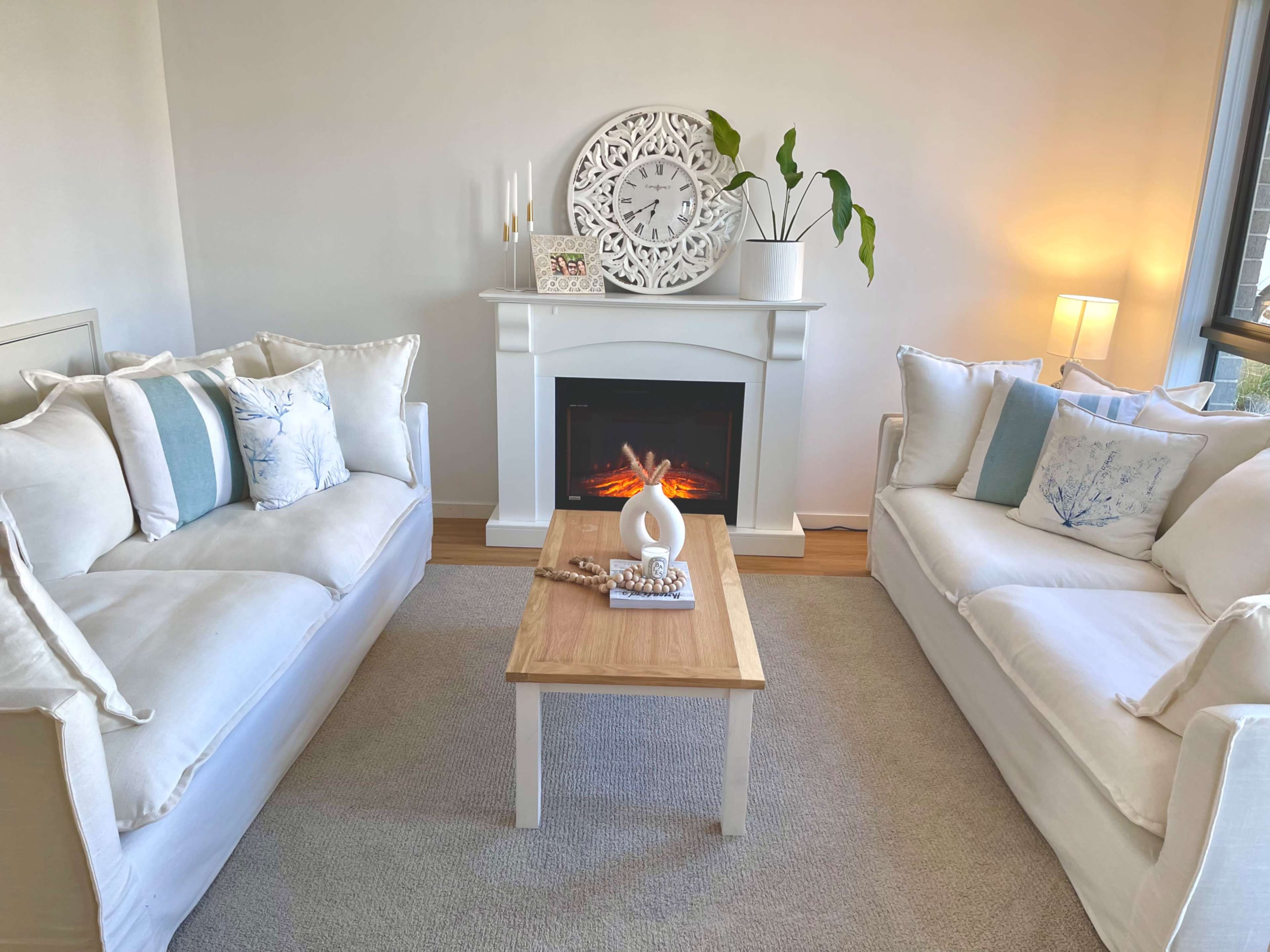 A cozy living room features two white sofas with decorative pillows, a wooden coffee table, and an electric fireplace beneath a wall clock and a potted plant.