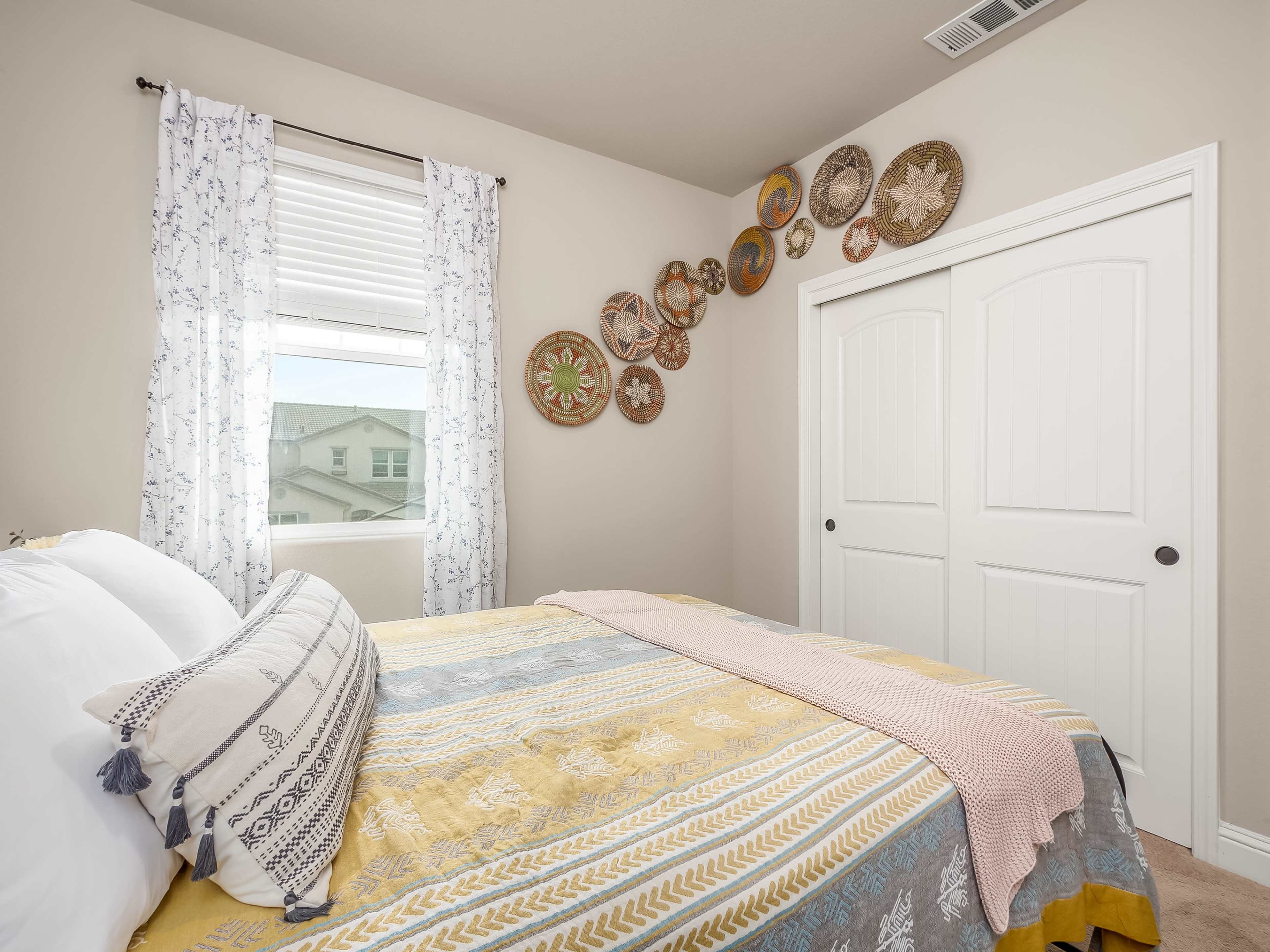 A cozy bedroom featuring a patterned bedspread, decorative pillows, and woven wall art above a window with sheer curtains.