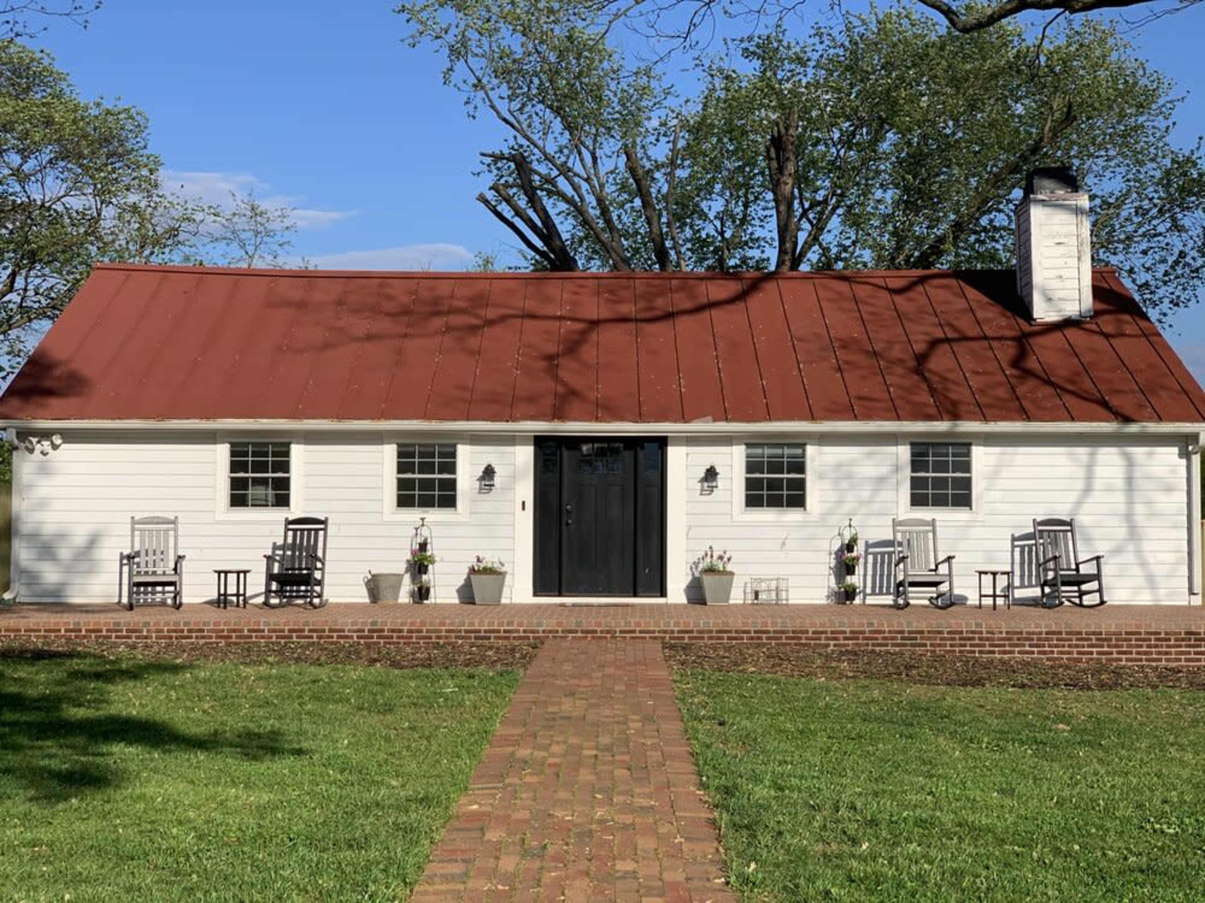 A white house with a red metal roof, a black front door, and a brick pathway leading to a covered porch with rocking chairs and potted plants.