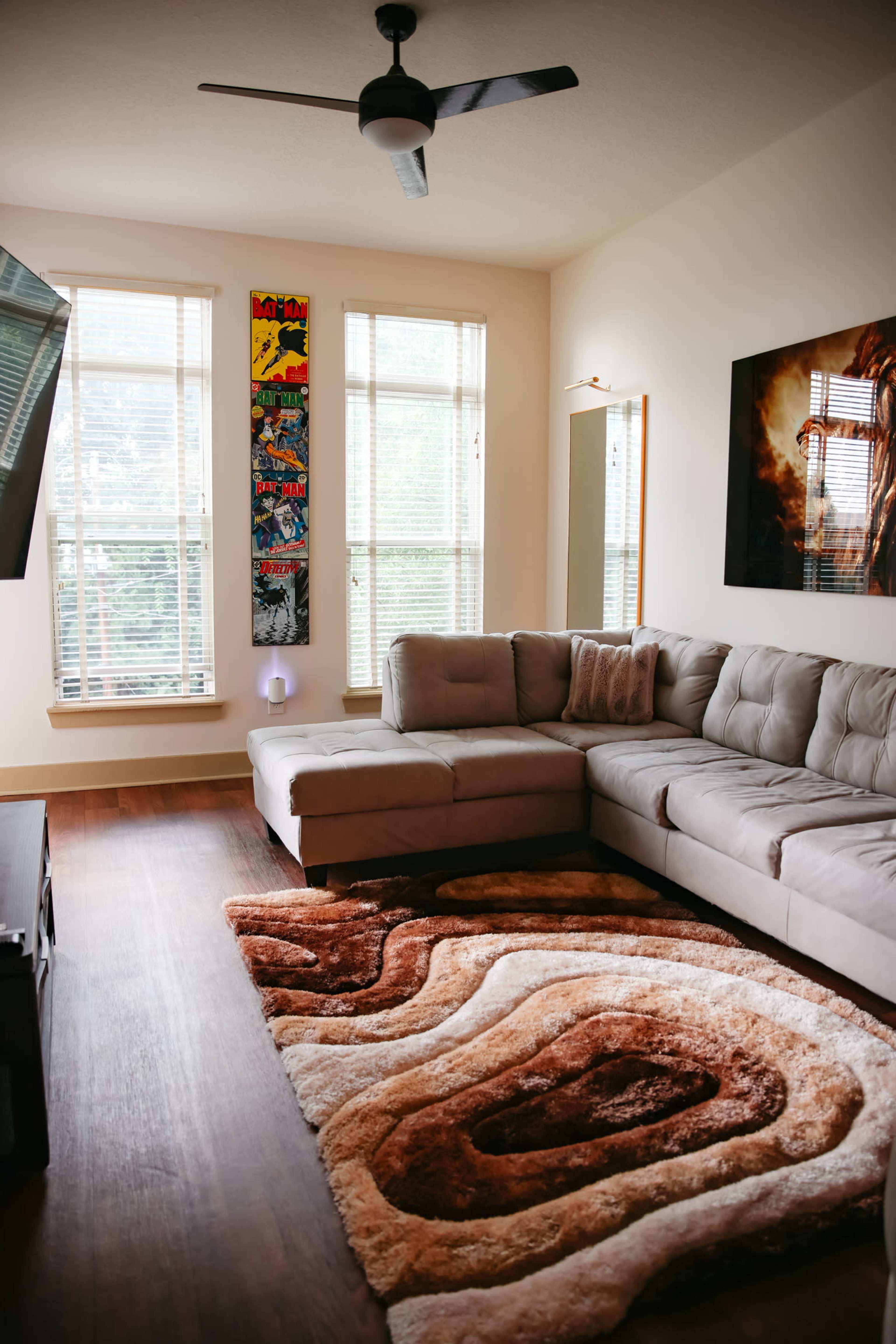 The image shows a modern living room with a sectional sofa, a patterned rug, and large windows allowing natural light.