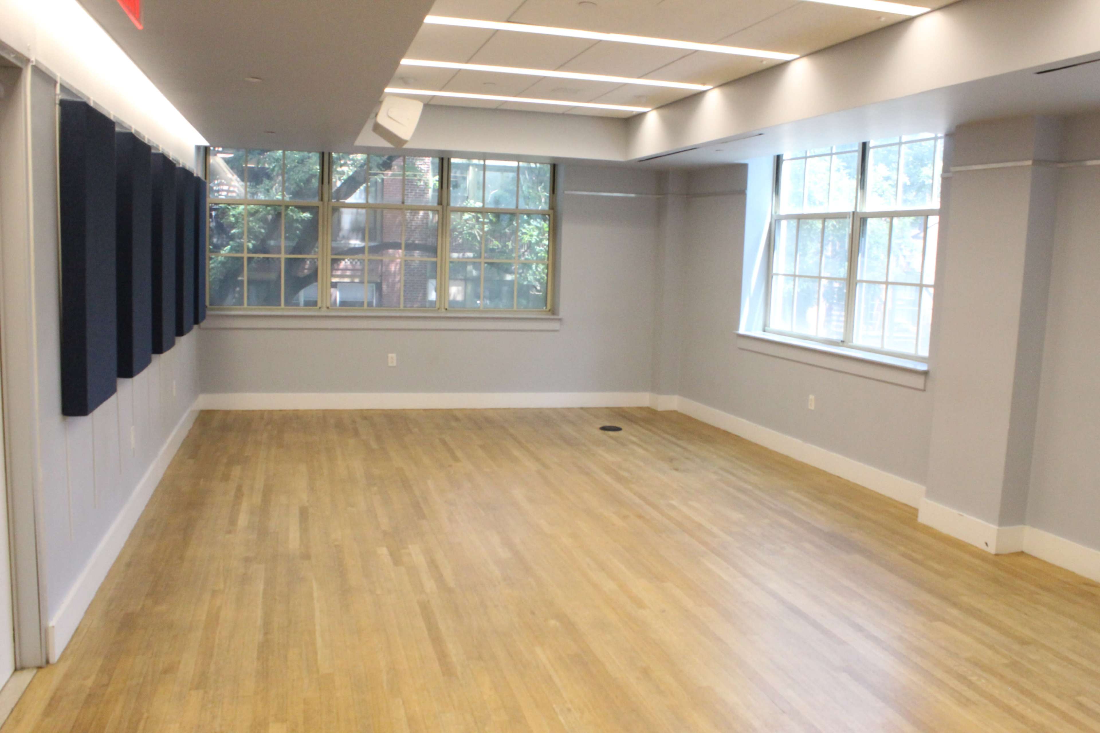 The image shows a spacious, empty room with wooden flooring and large windows providing natural light.