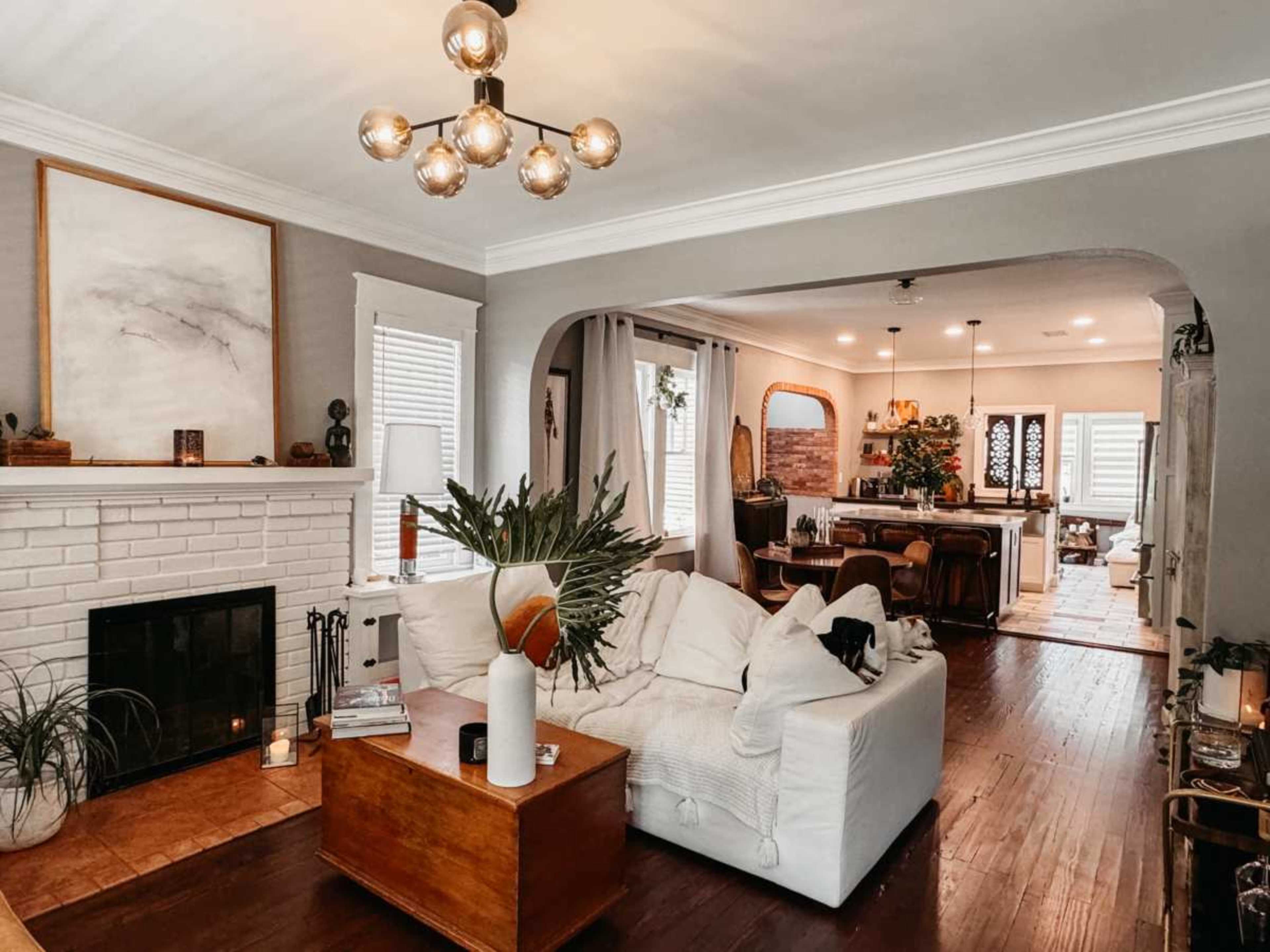 The image shows a cozy living room with a white sofa, a wooden coffee table, and a fireplace, leading to a dining area in the background.