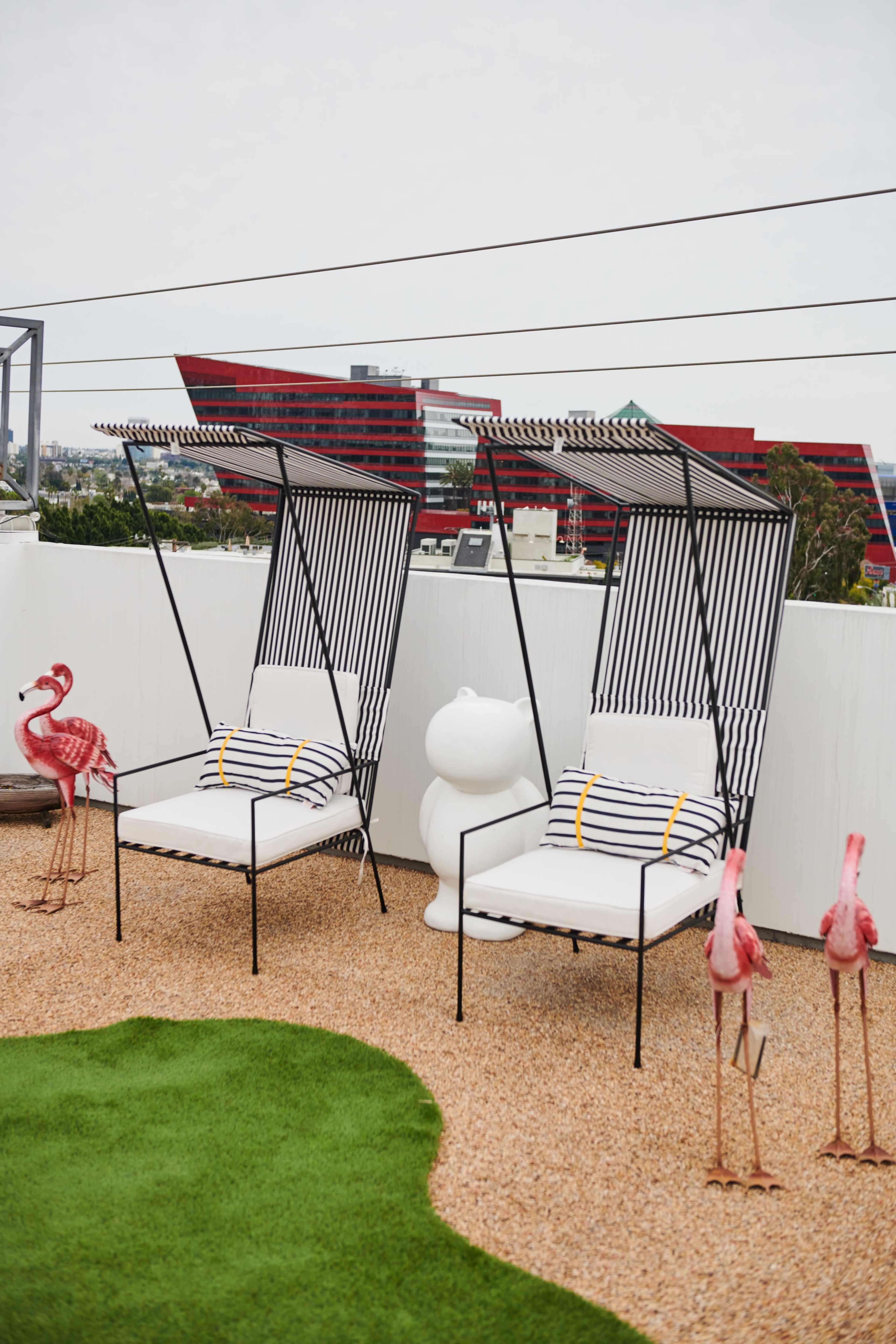 Two striped lounge chairs with canopies sit on a gravel patio surrounded by decorative flamingos and a green lawn, with modern buildings in the background.