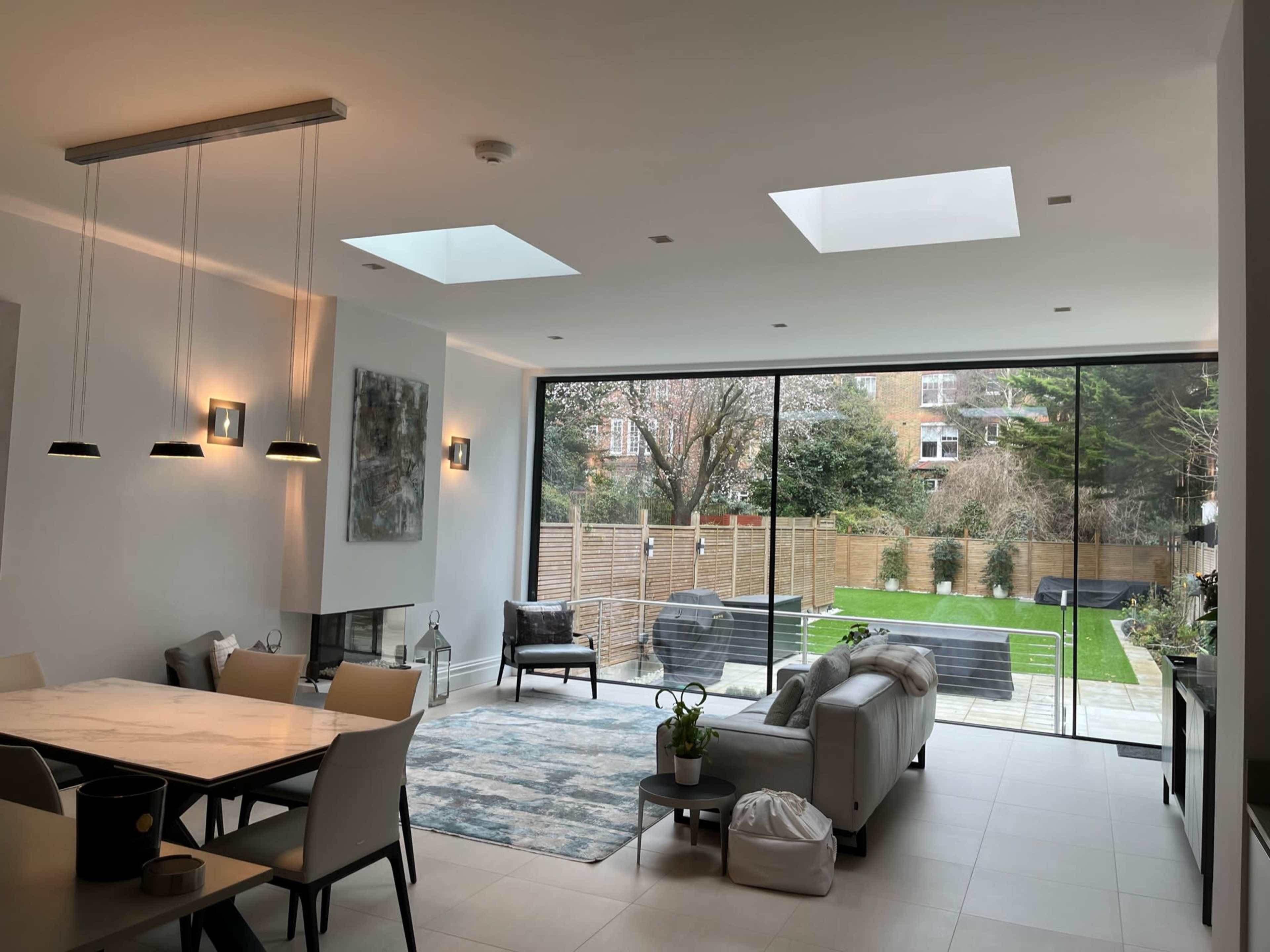 A modern living room with large glass doors leading to a garden, featuring a dining area, a couch, and a rug.