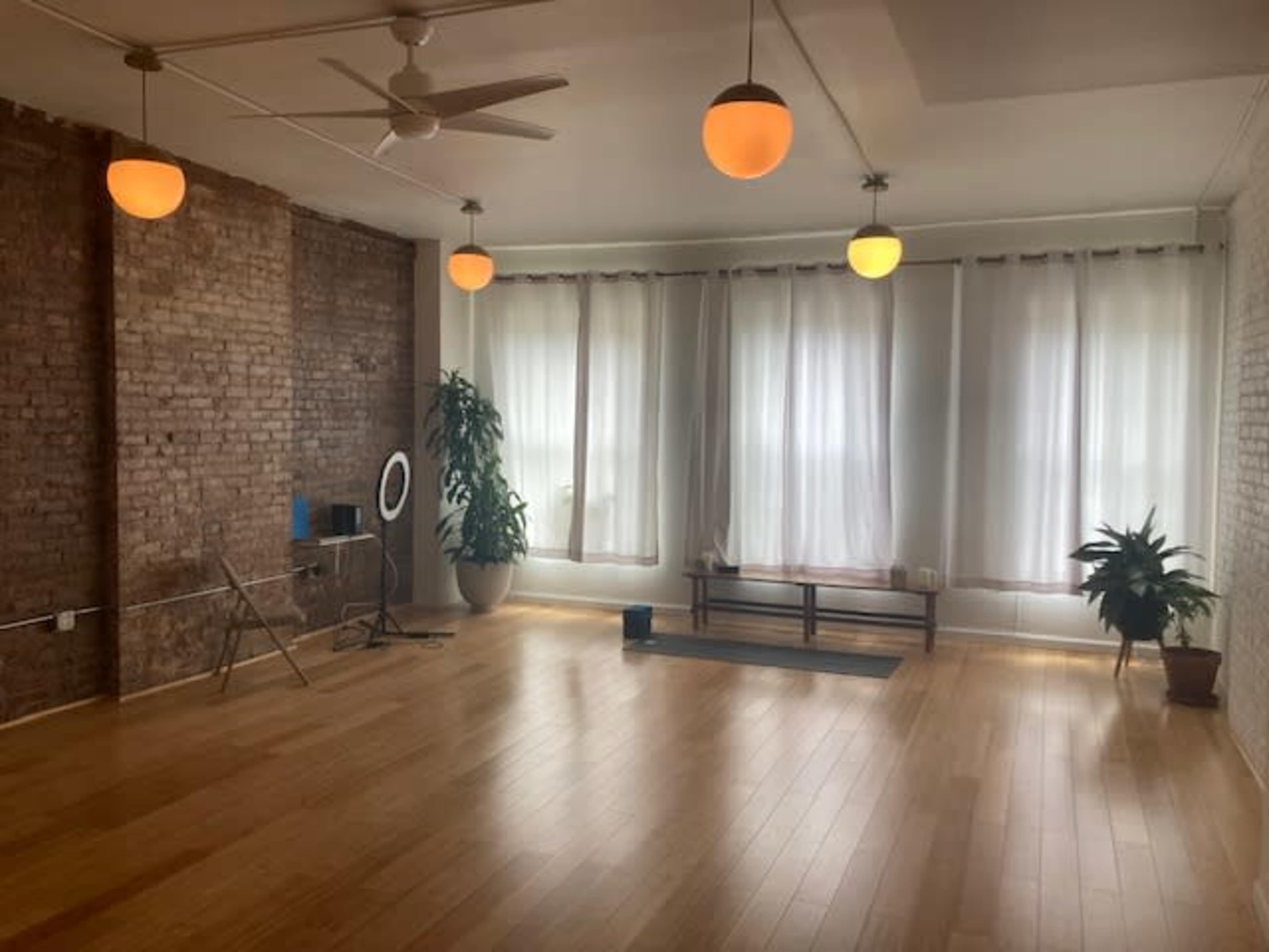 A bright, spacious room with wooden floors, large windows covered by sheer white curtains, and warm pendant lights hanging from the ceiling.