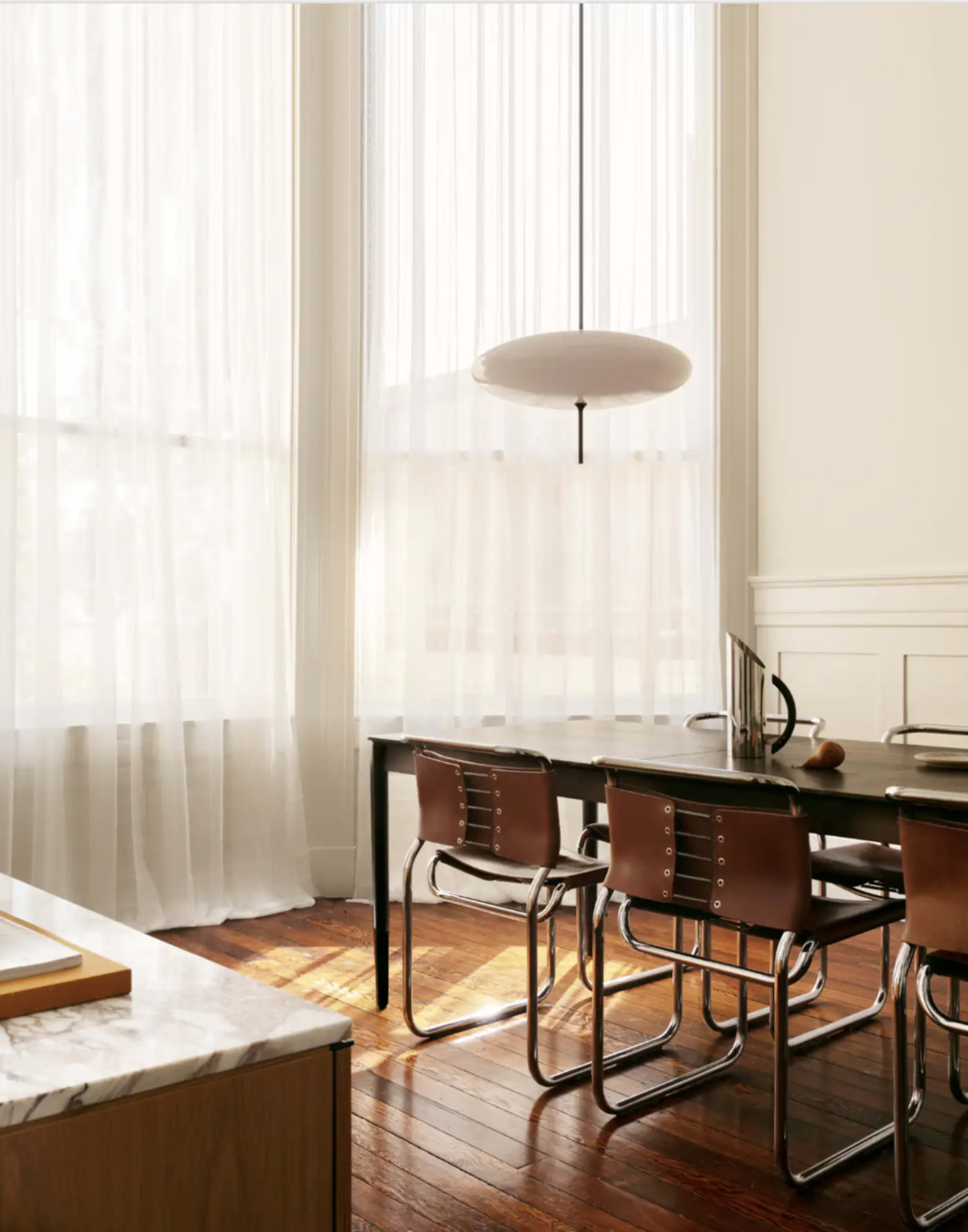 A dining area features a wooden table surrounded by metal-framed chairs, illuminated by natural light through sheer curtains.