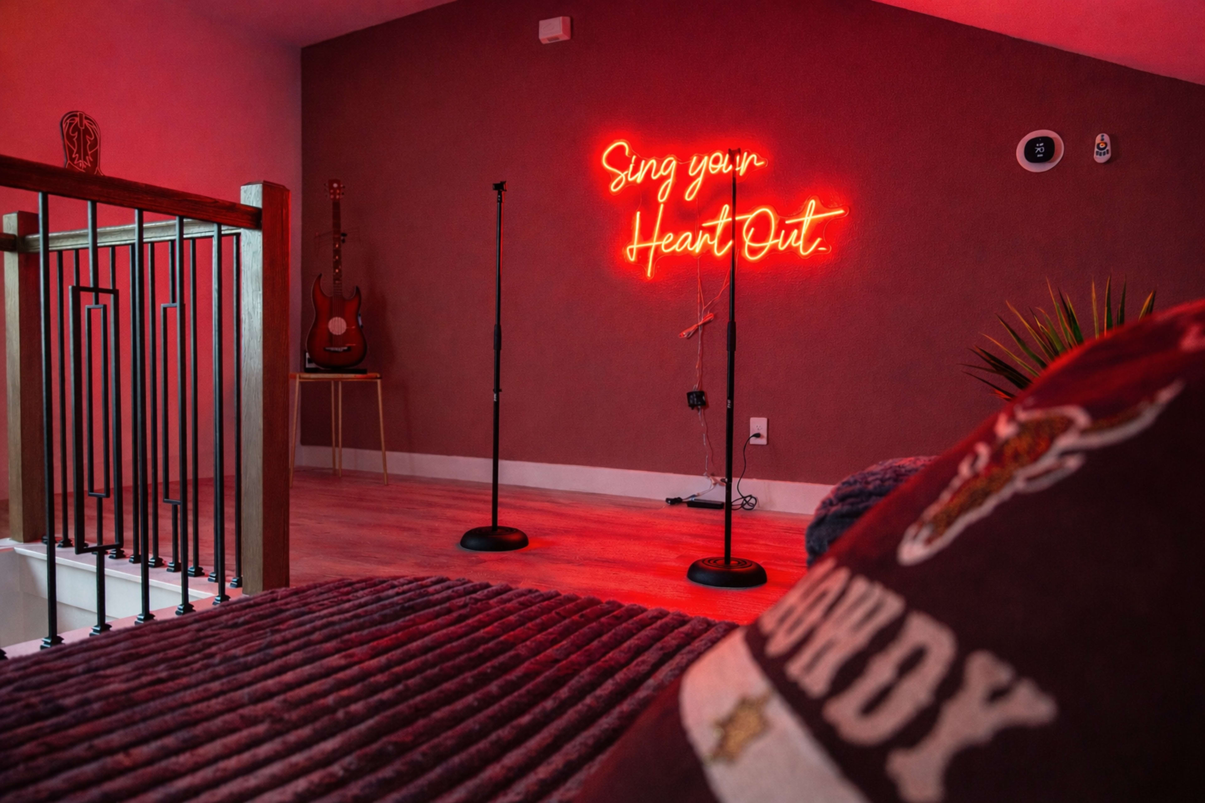 The image shows a cozy room with a red neon sign reading "Sing your Heart Out," two microphone stands, and an acoustic guitar on display.