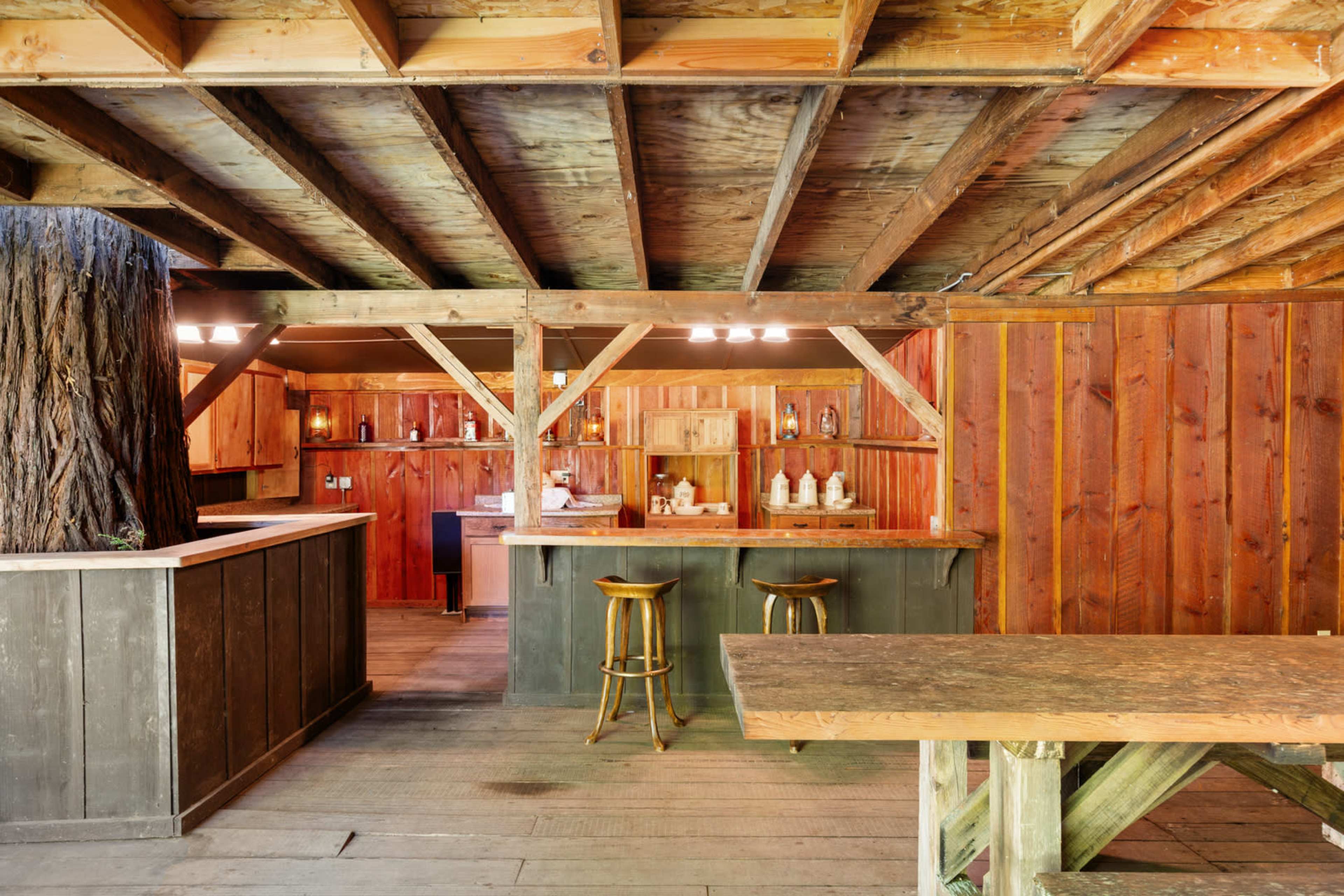 The image shows a rustic interior of a cabin featuring wooden beams, a central tree trunk, a bar area with stools, and shelves lined with bottles.