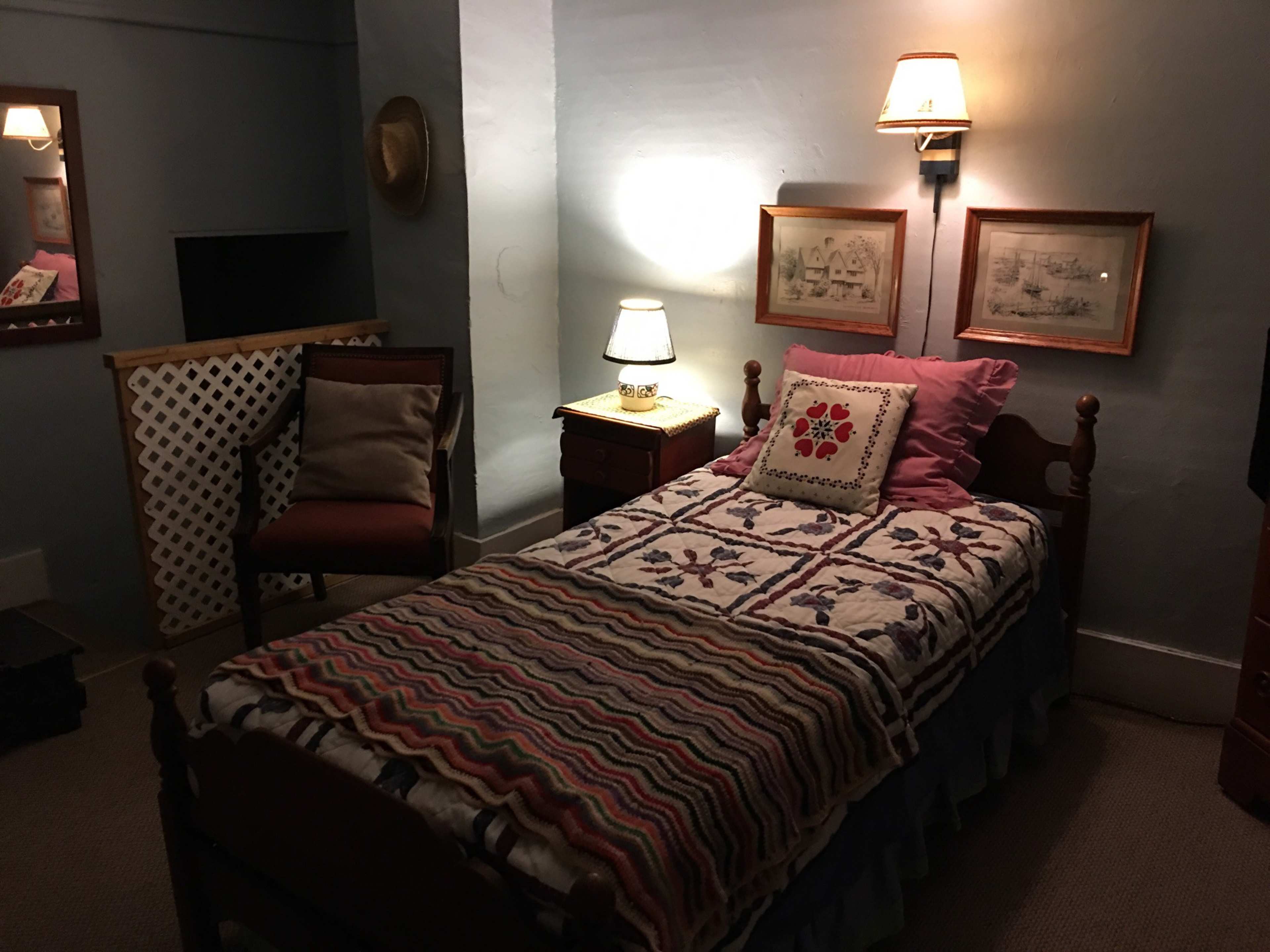 The image depicts a small bedroom featuring a single bed with a patterned quilt, a chair, a nightstand with a lamp, and framed pictures on the wall.