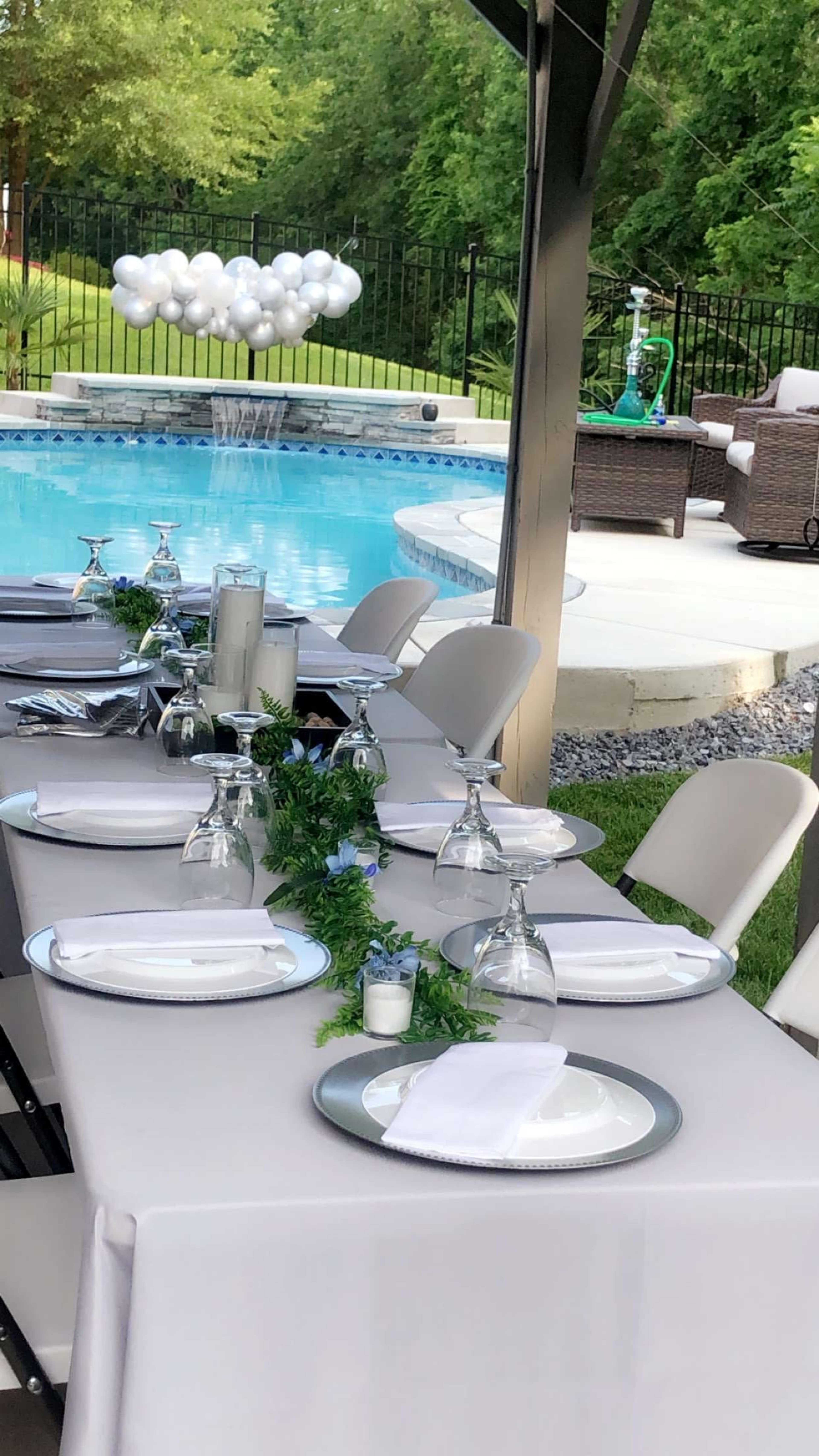 A neatly arranged outdoor dining table is set beside a swimming pool, with decorative greenery and glassware, and a cluster of balloons in the background.