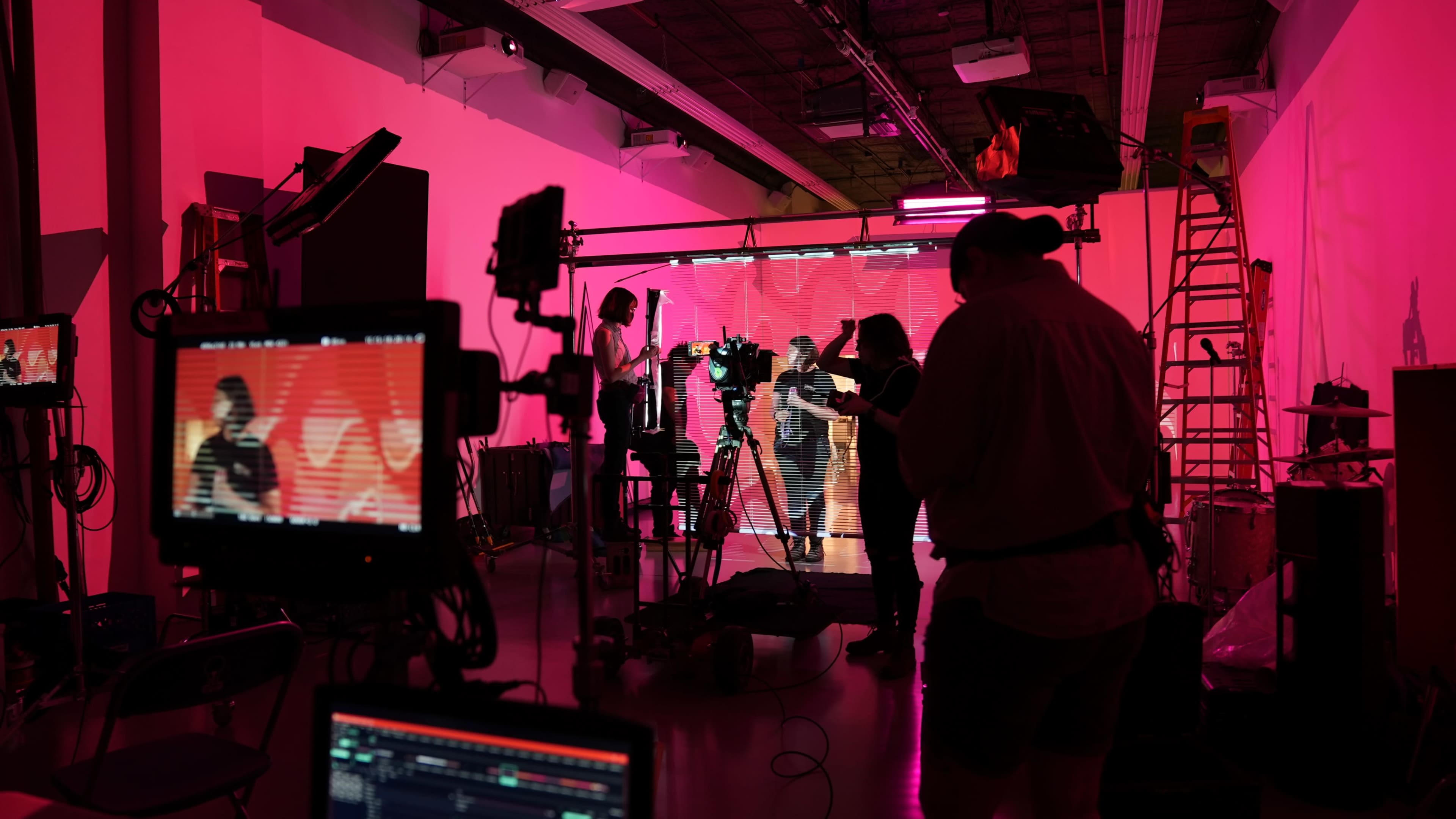 A film crew sets up equipment on a brightly lit set with pink lighting and backdrops.