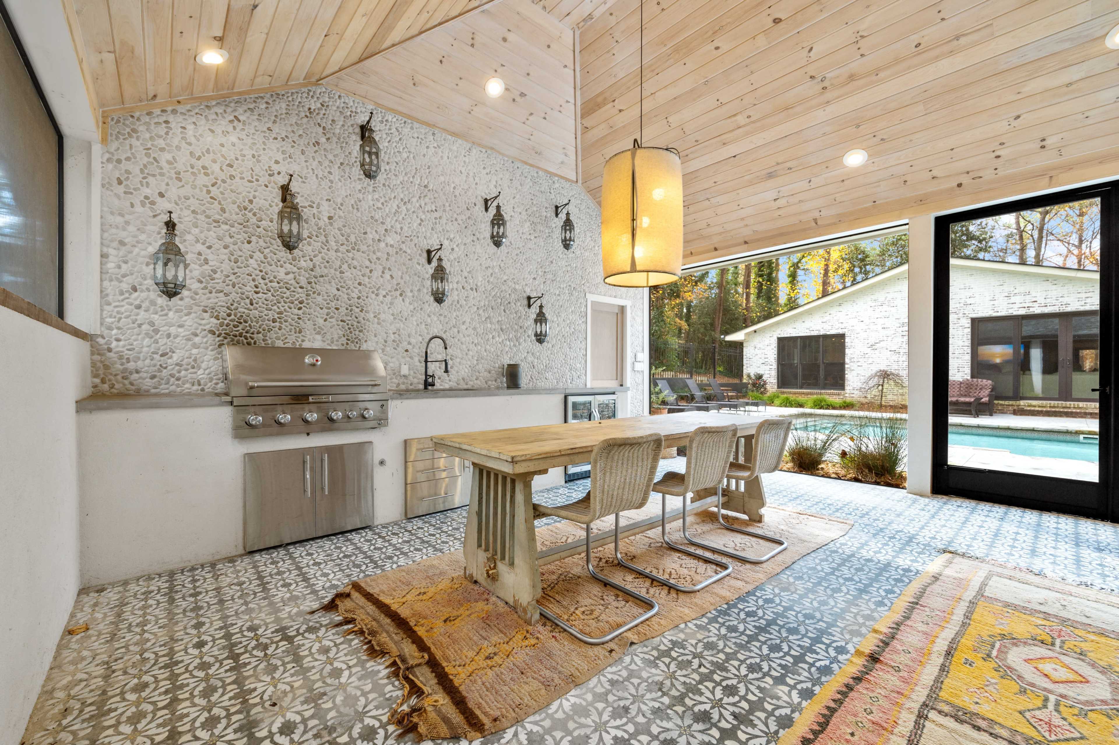 The image shows a modern outdoor kitchen area featuring a stainless steel grill, a wooden dining table with metal chairs, and decorative tile flooring.