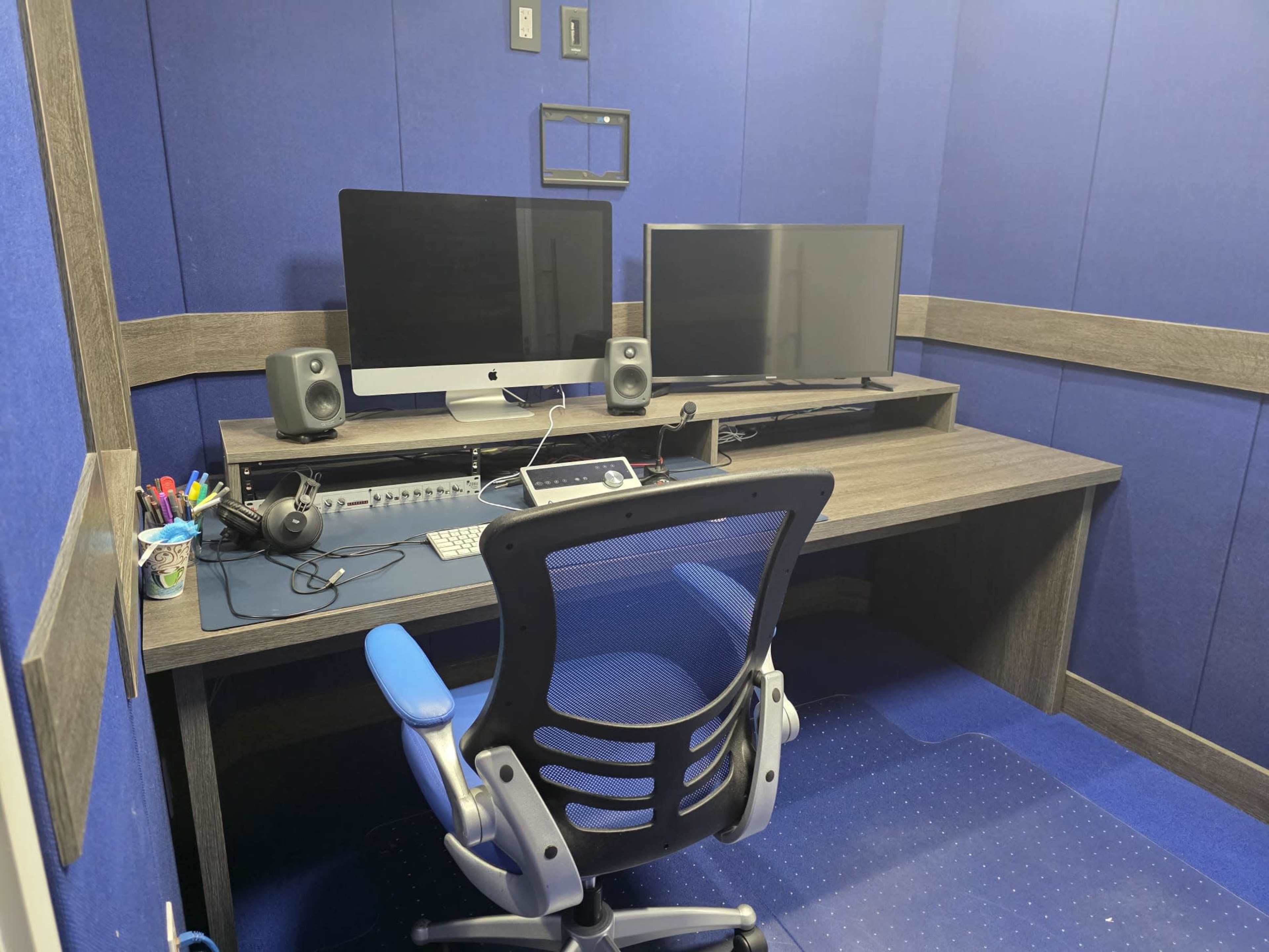 The image shows a small office booth with a wooden desk, two monitors, speakers, and a blue ergonomic chair on a blue carpeted floor.