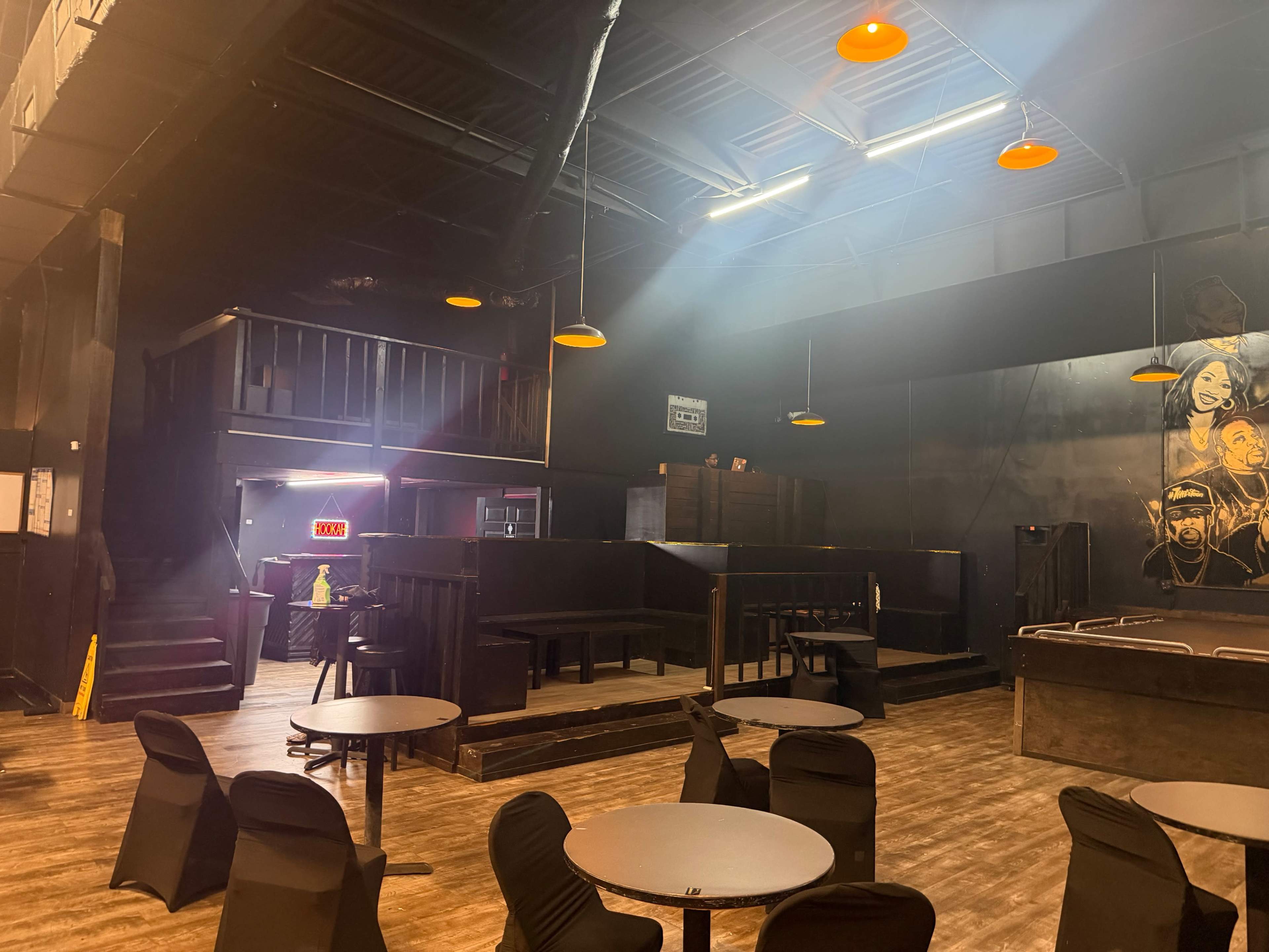The image shows the interior of a dimly lit event space featuring black walls, scattered round tables with chairs, and orange pendant lights hanging from the ceiling.