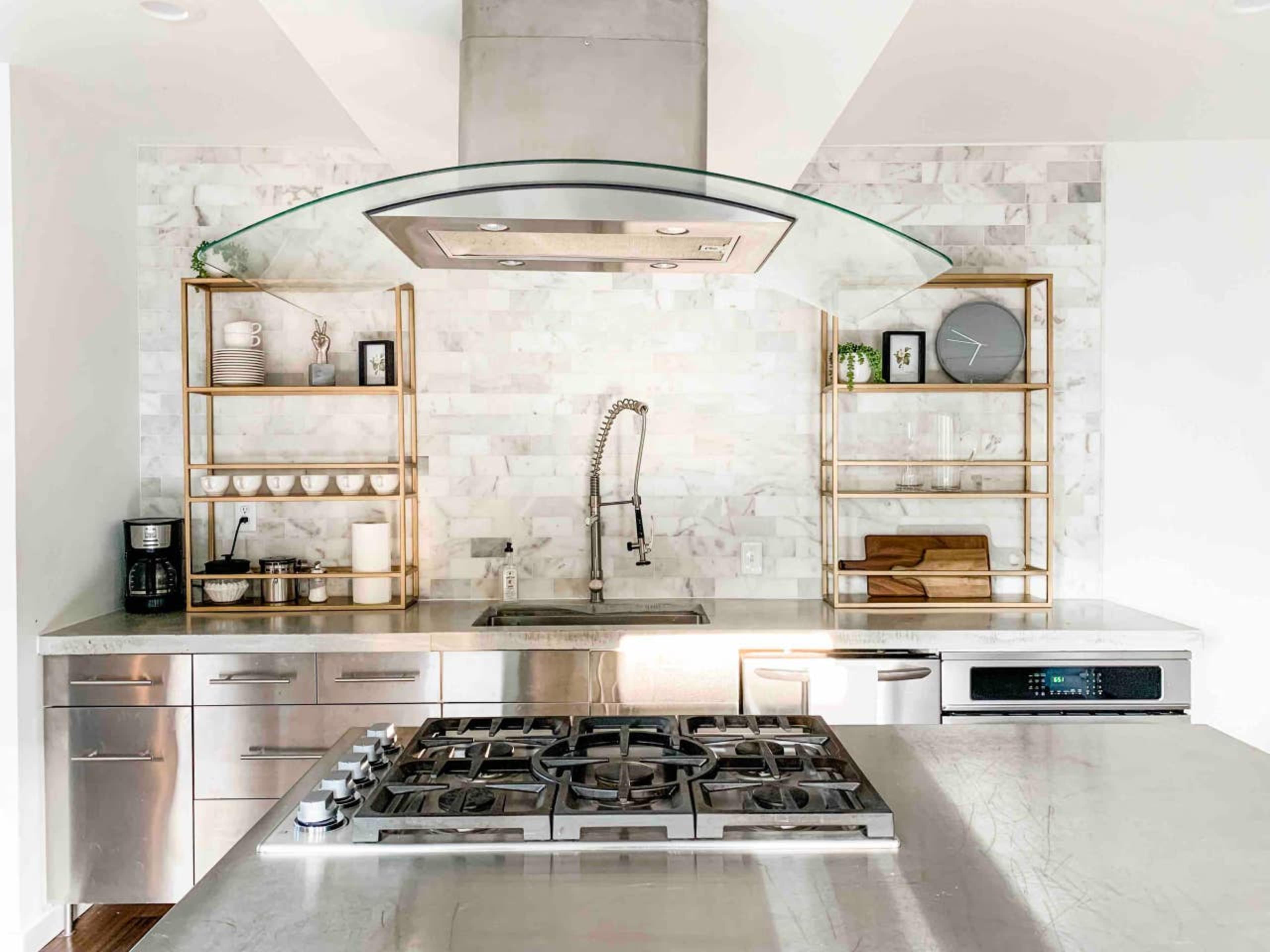 A modern kitchen featuring stainless steel appliances, a gas stovetop, a marble backsplash, and open metal shelving displaying dishware and utensils.